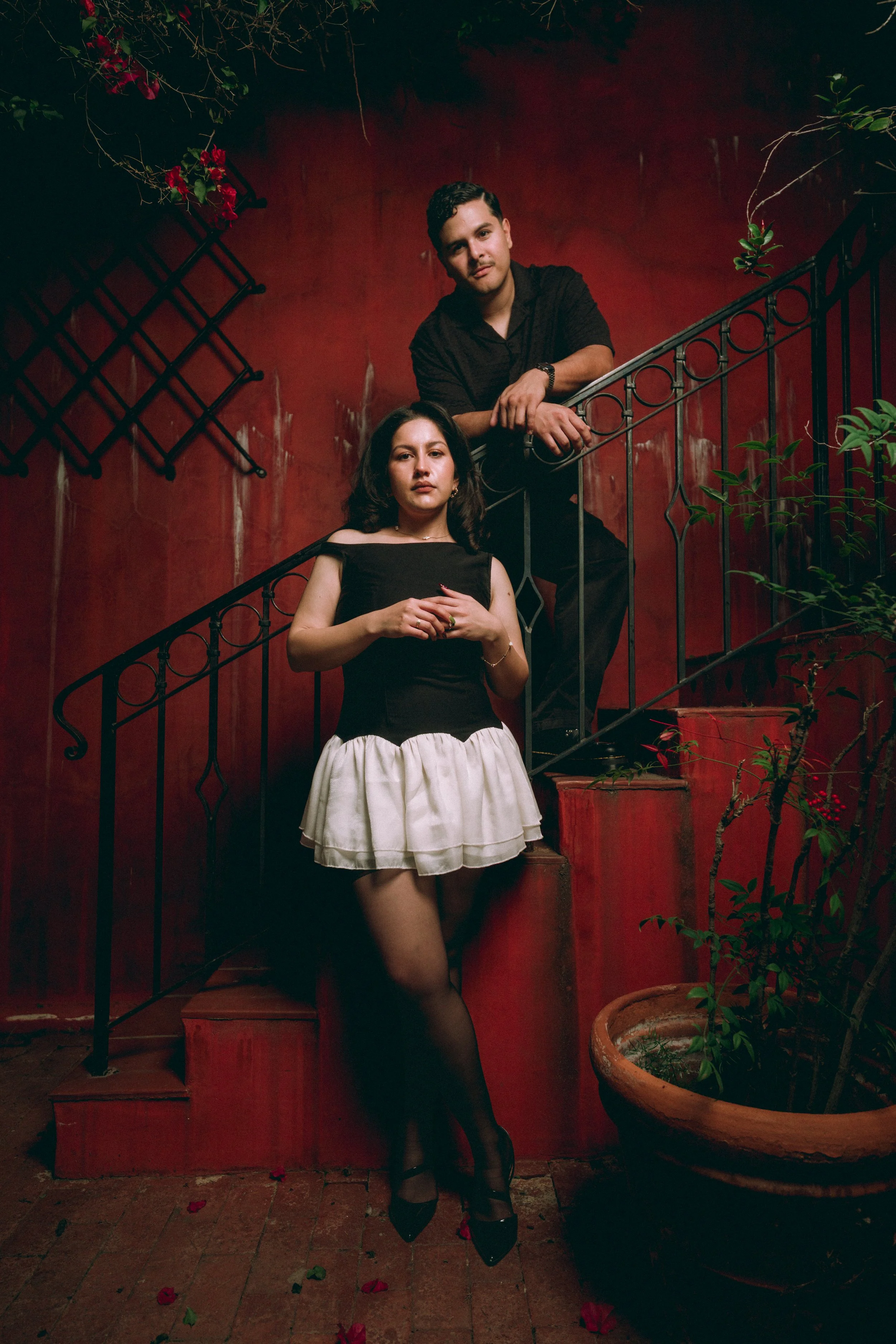 A young man and woman are standing on a staircase with a red wall background, surrounded by plants and flowers. The woman is wearing a black top and a white skirt, and has dark hair. The man is leaning on the railing, wearing a black shirt, and has c
