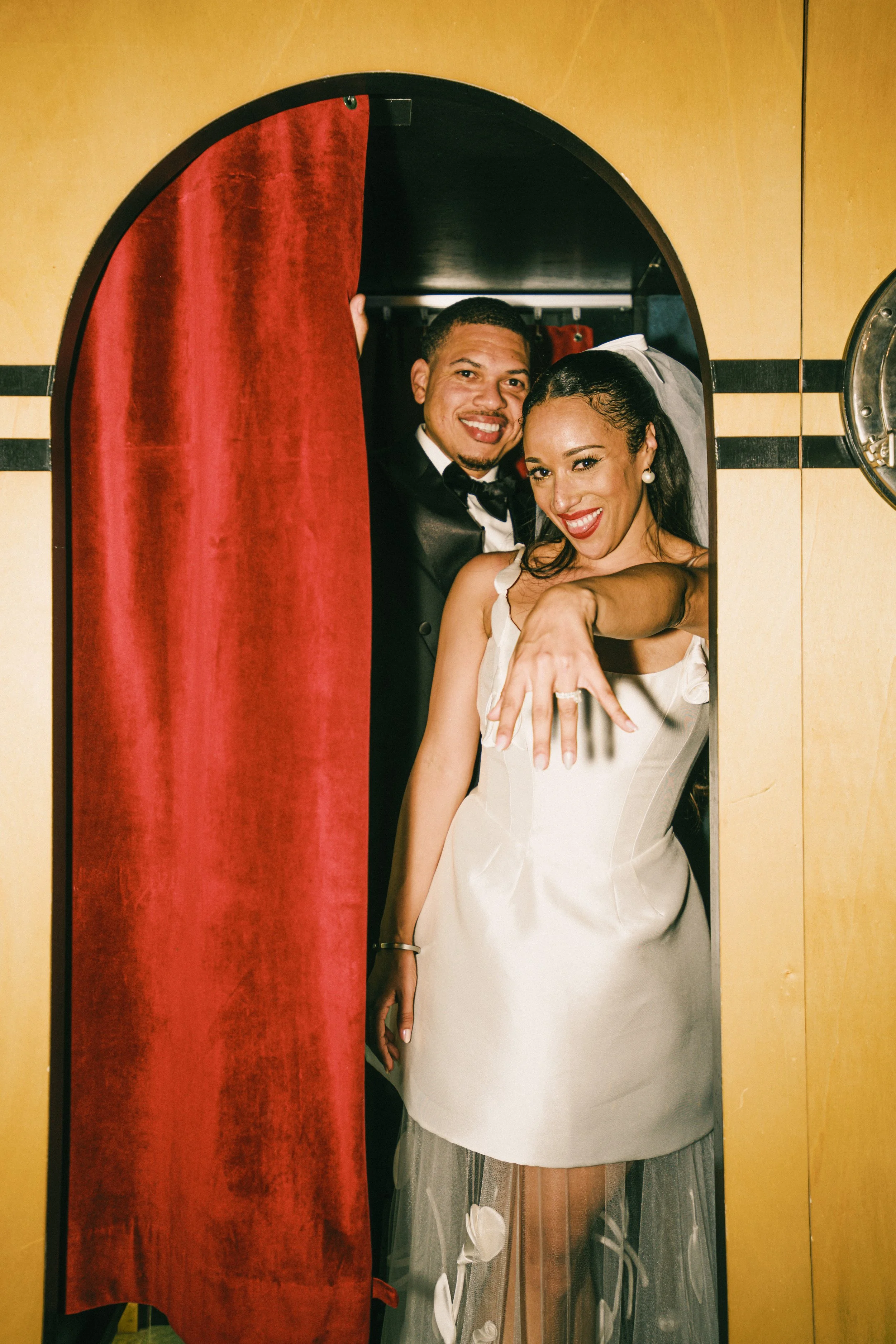 A bride and groom smile as they emerge through a porthole-like opening with a red velvet curtain. The bride is wearing a white wedding dress with her hair styled in loose waves, and the groom is dressed in a black tuxedo. They are both pointing forwa