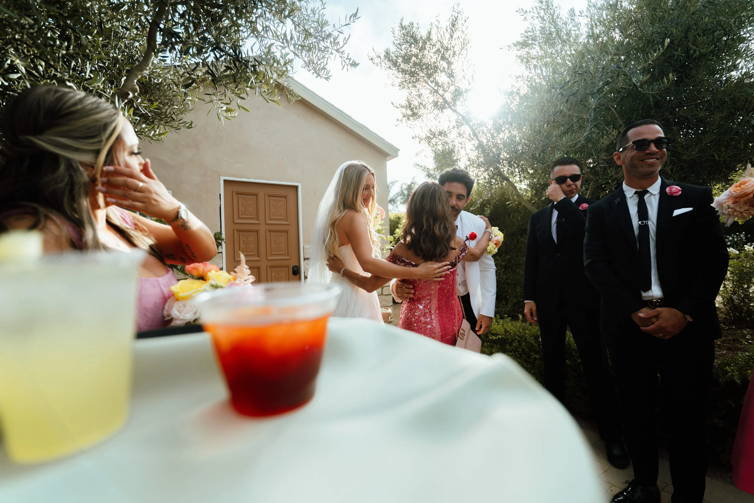 A group of people at a wedding reception outdoors, including women in dresses and men in suits with sunglasses, standing near a table with drinks and flowers, with a house and trees in the background.