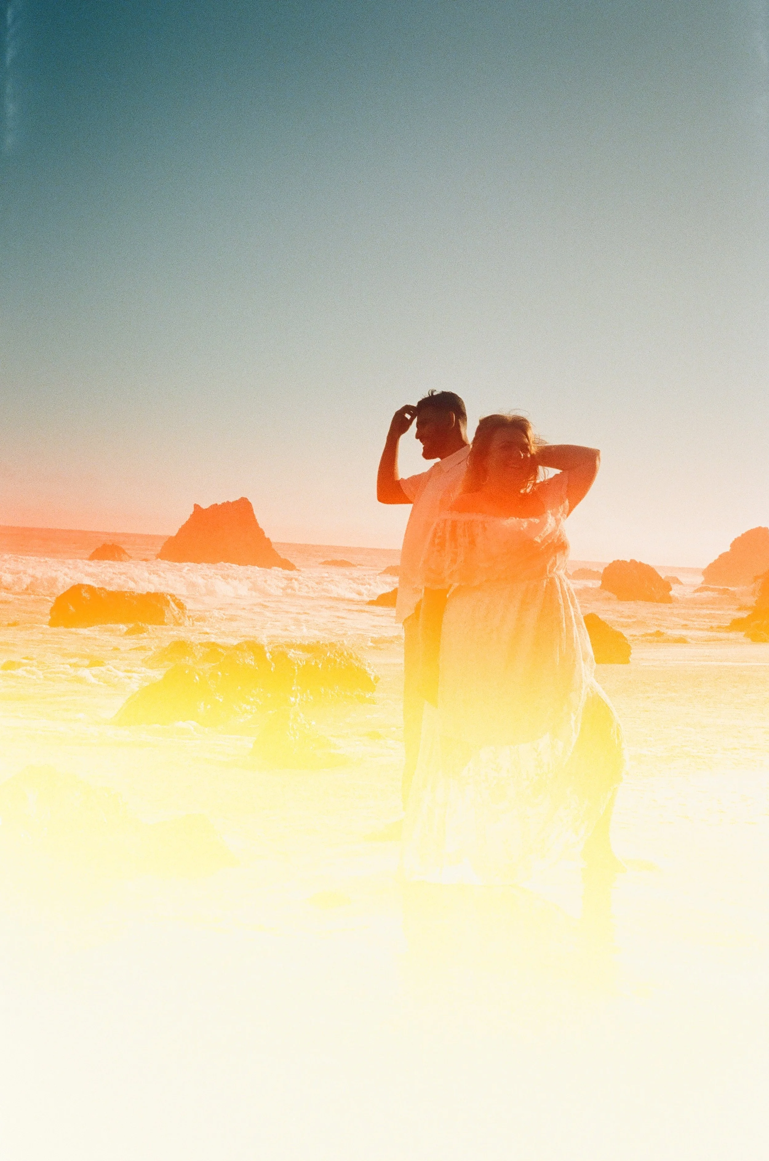 A man and woman standing on the beach at sunset with rocks in the background.