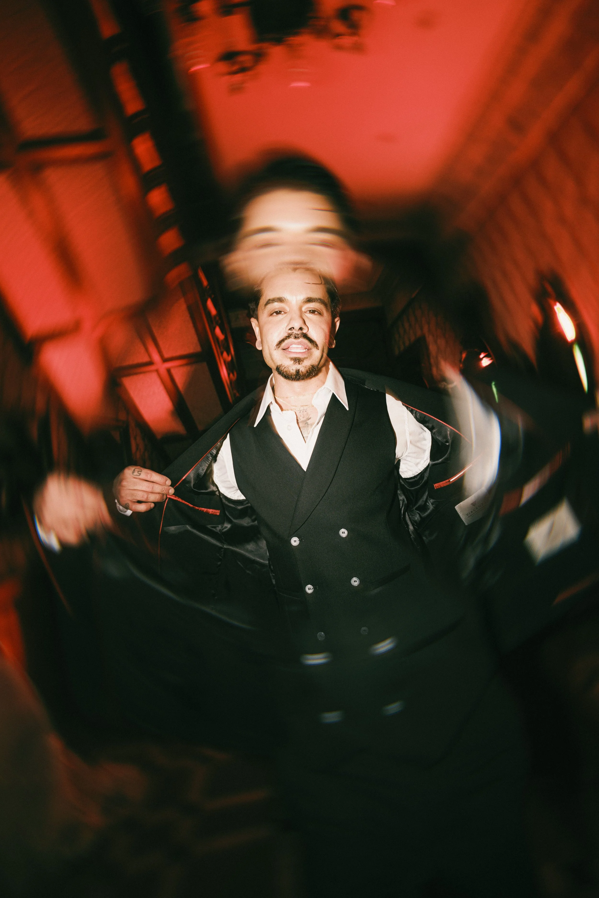 A man with a mustache and beard looking into a camera with a distorted, fisheye effect. He is wearing a black vest over a white shirt and is holding the front of the vest open with one hand. The background is blurred with warm, reddish lighting.
