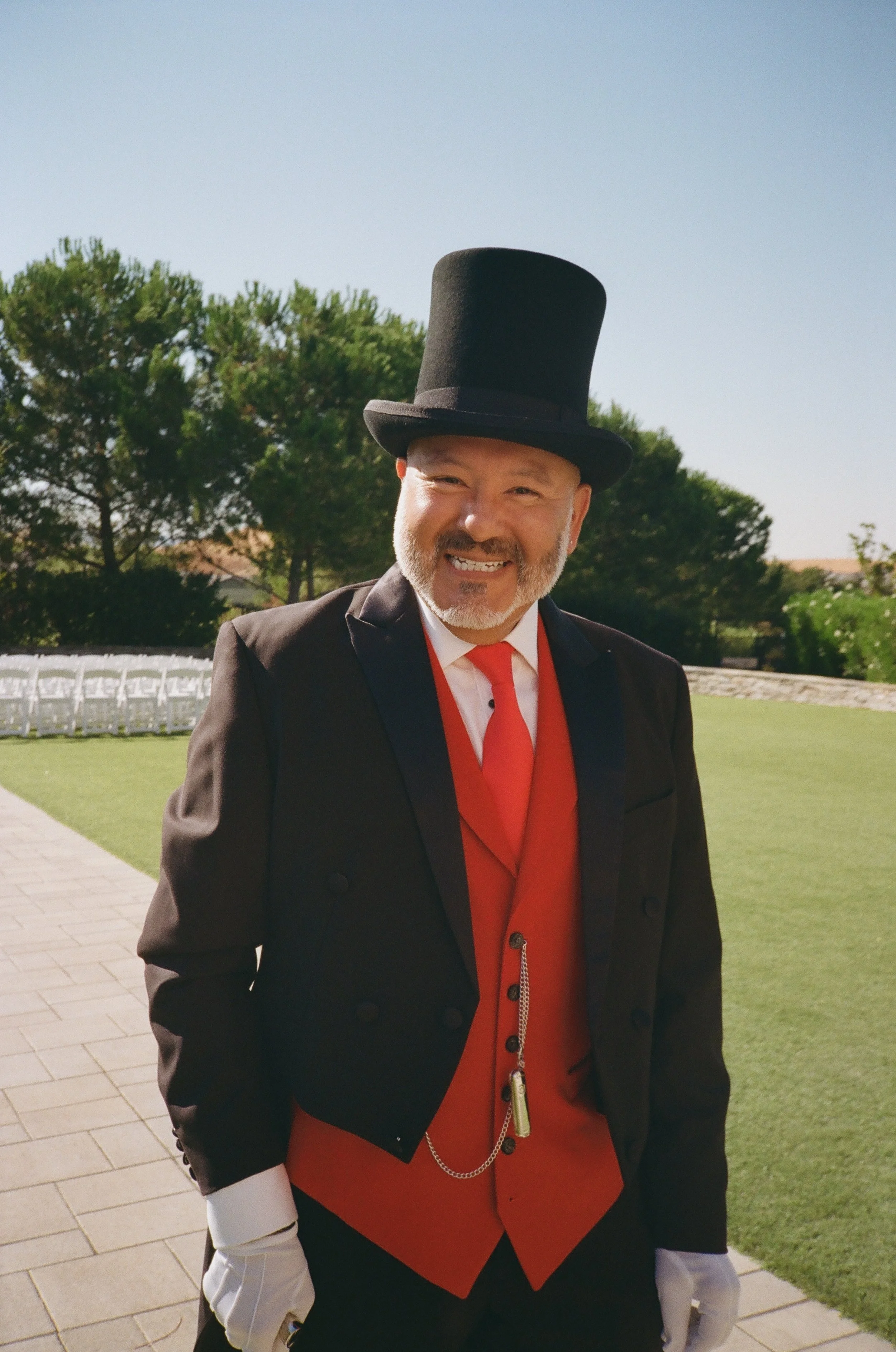 A man dressed as a ringmaster, wearing a black top hat, black tailcoat, red vest, red tie, white gloves, and a pocket watch chain, smiling outdoors on a sunny day with a grassy area and trees in the background.