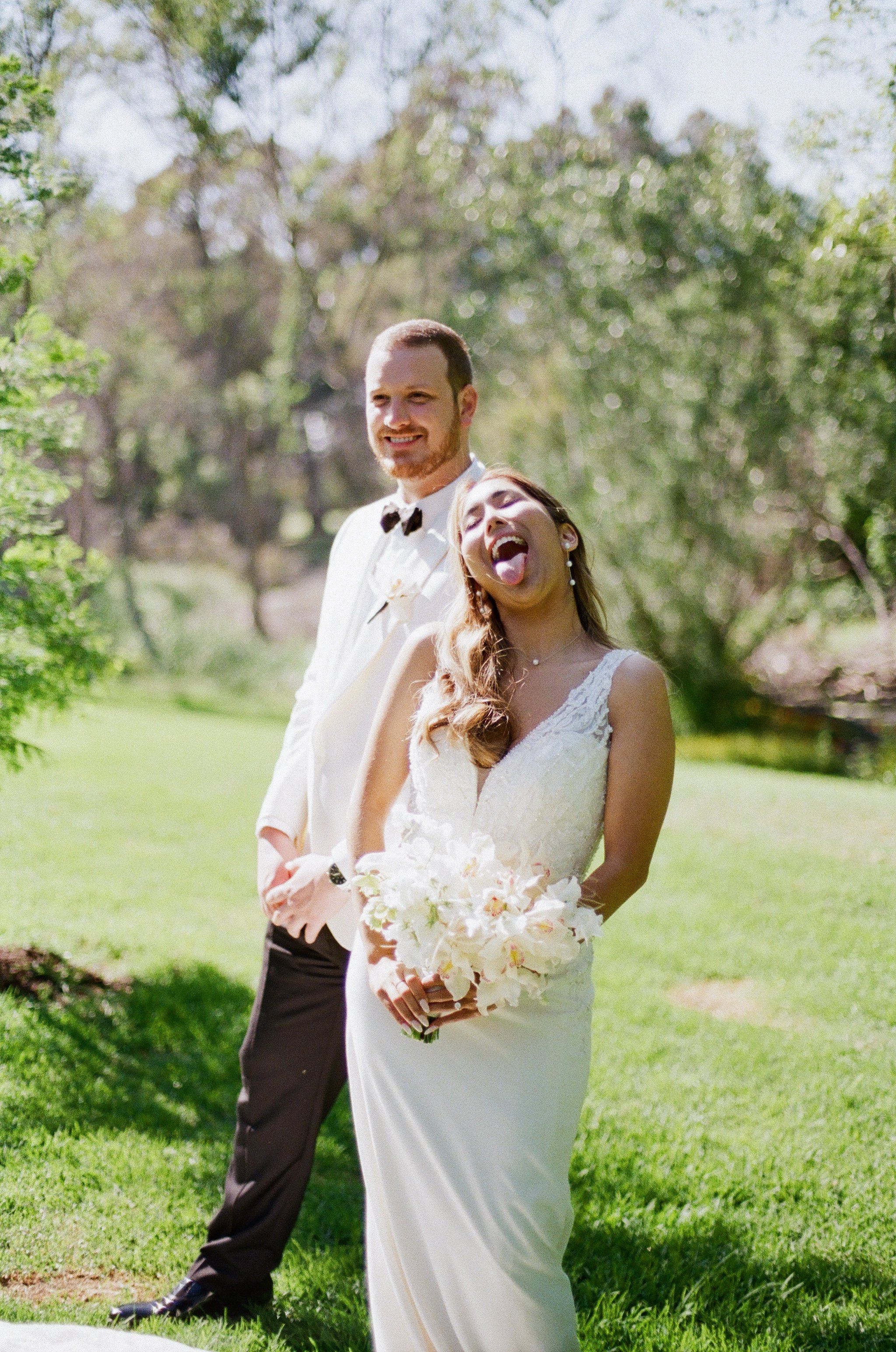 A bride holding a bouquet of white flowers stands outdoors on a grassy lawn, smiling, with a groom in a white suit and bow tie standing behind her in an outdoor setting with trees and sunlight.