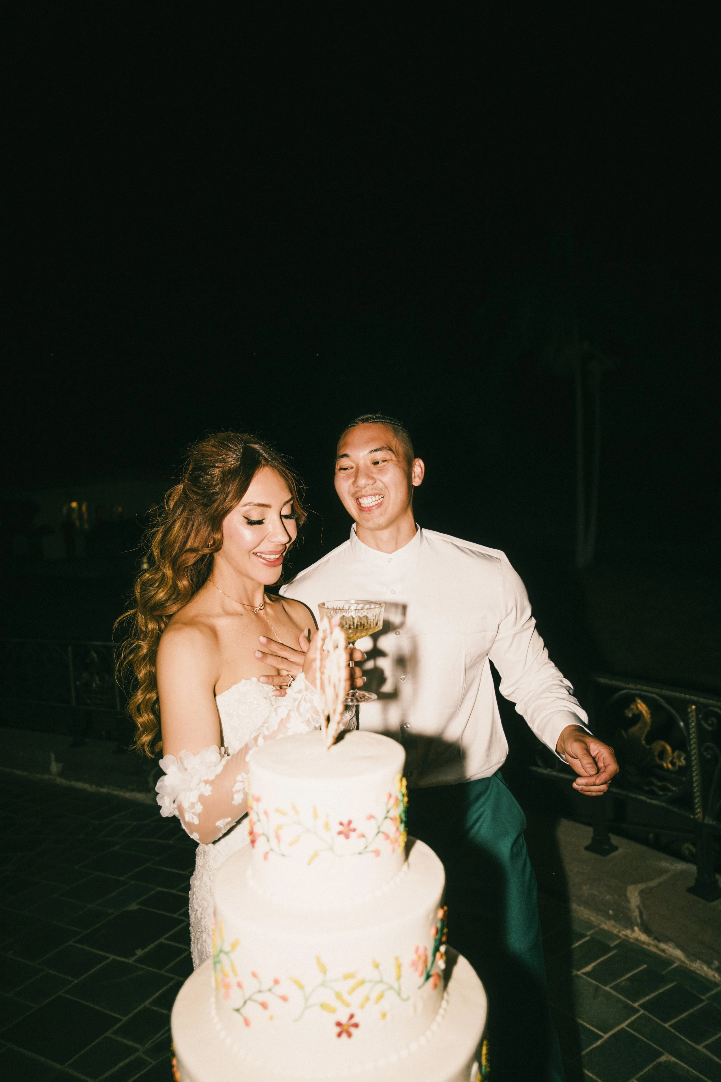 A bride and groom celebrating at their wedding reception near a decorated cake with floral designs, with a dark night sky in the background.
