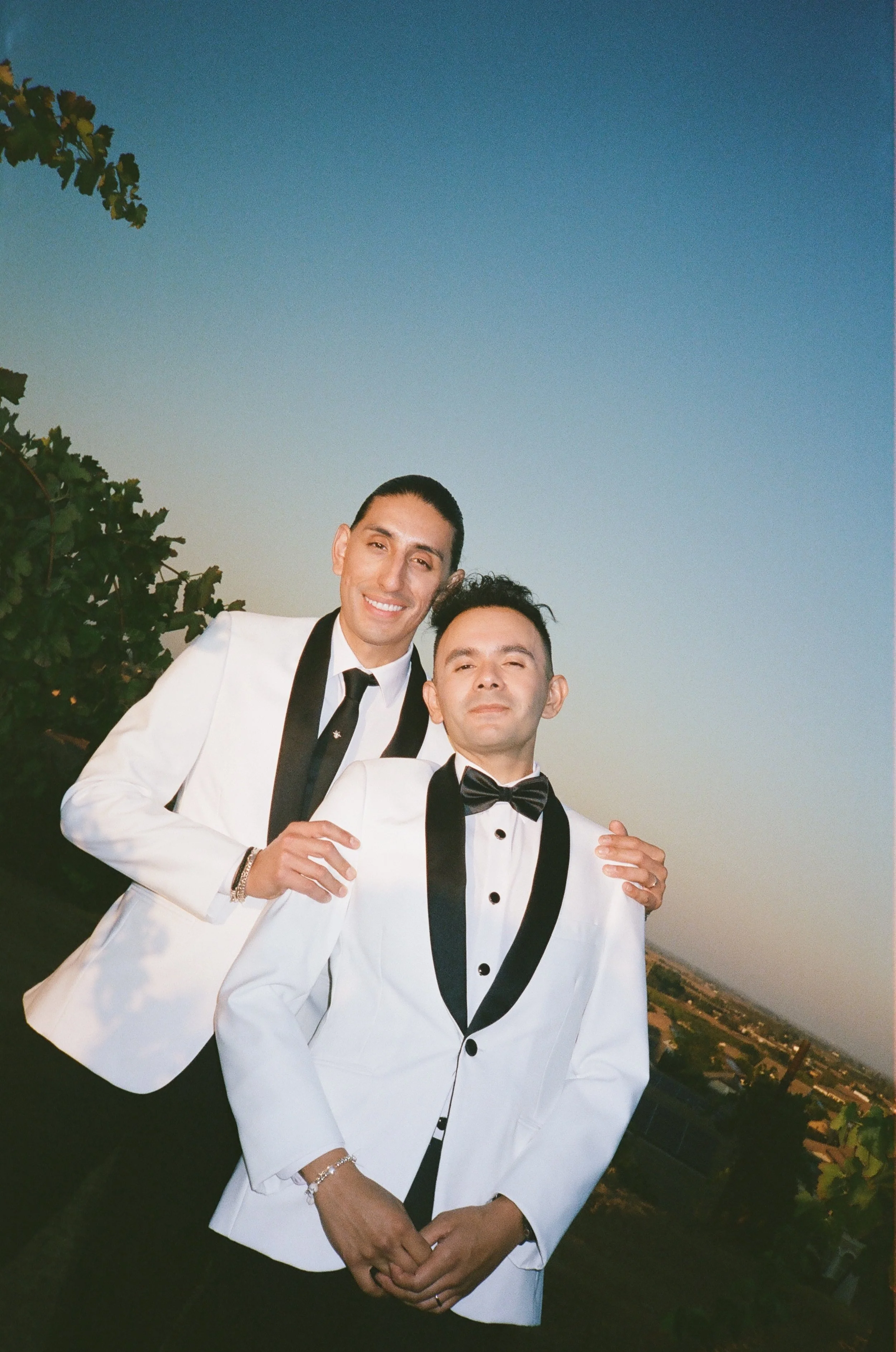 Two men dressed in white tuxedos with black lapels and bow ties, standing outdoors during sunset with a landscape in the background.