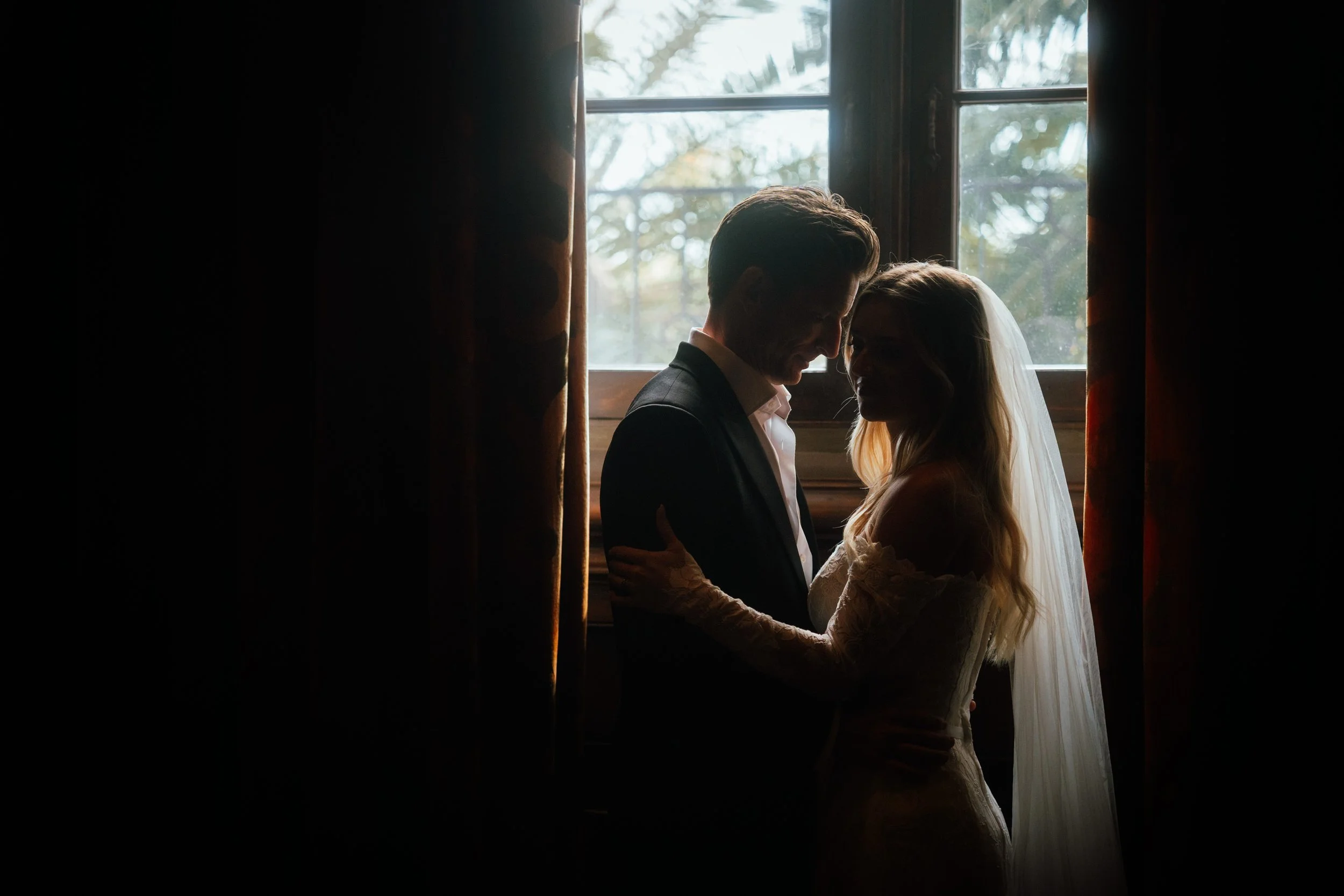 A couple dressed in wedding attire standing close together by a window, with sunlight shining through, creating a silhouette effect.