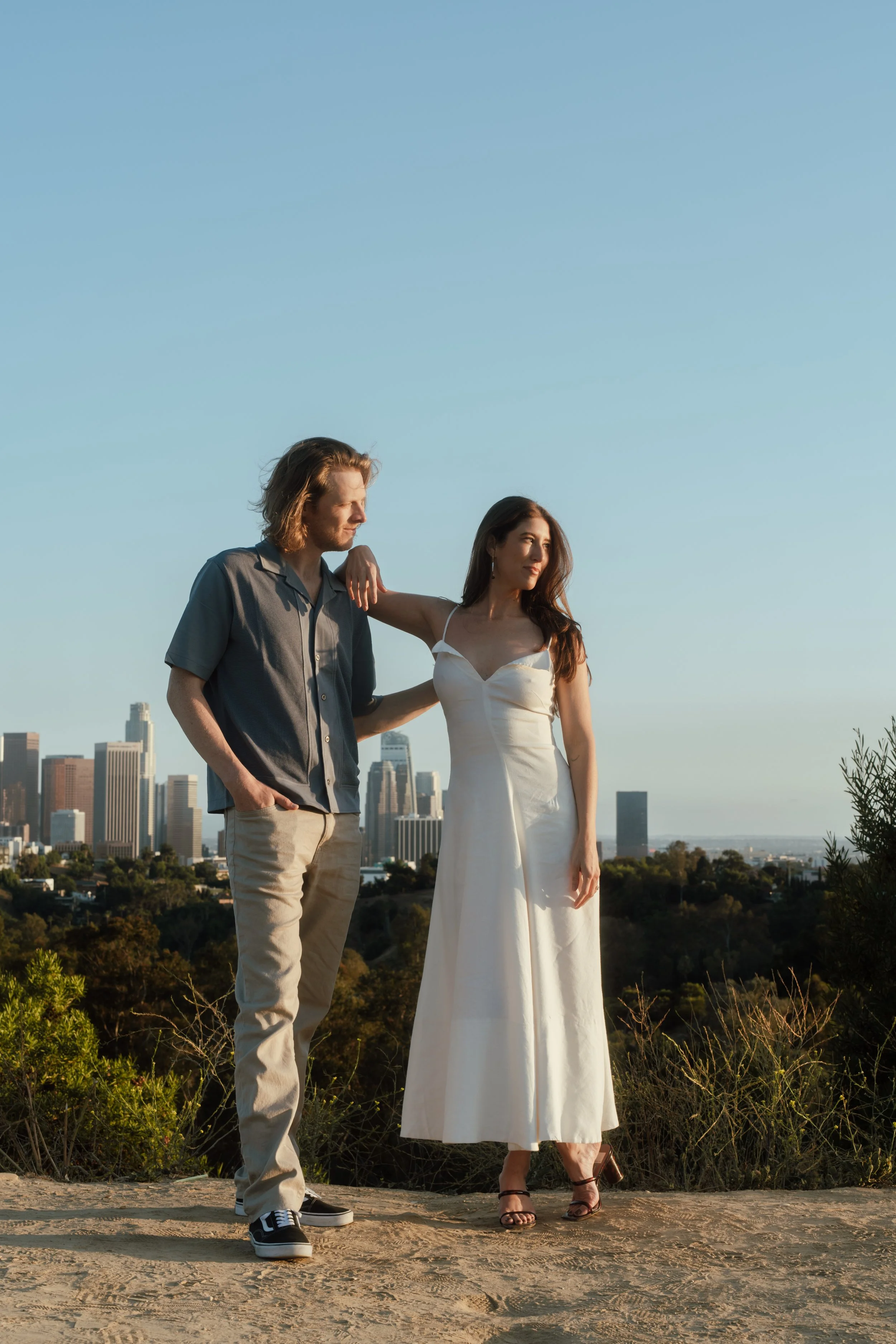 A man with shoulder-length hair and a woman in a white dress standing on a hill with a city skyline in the background during sunset.