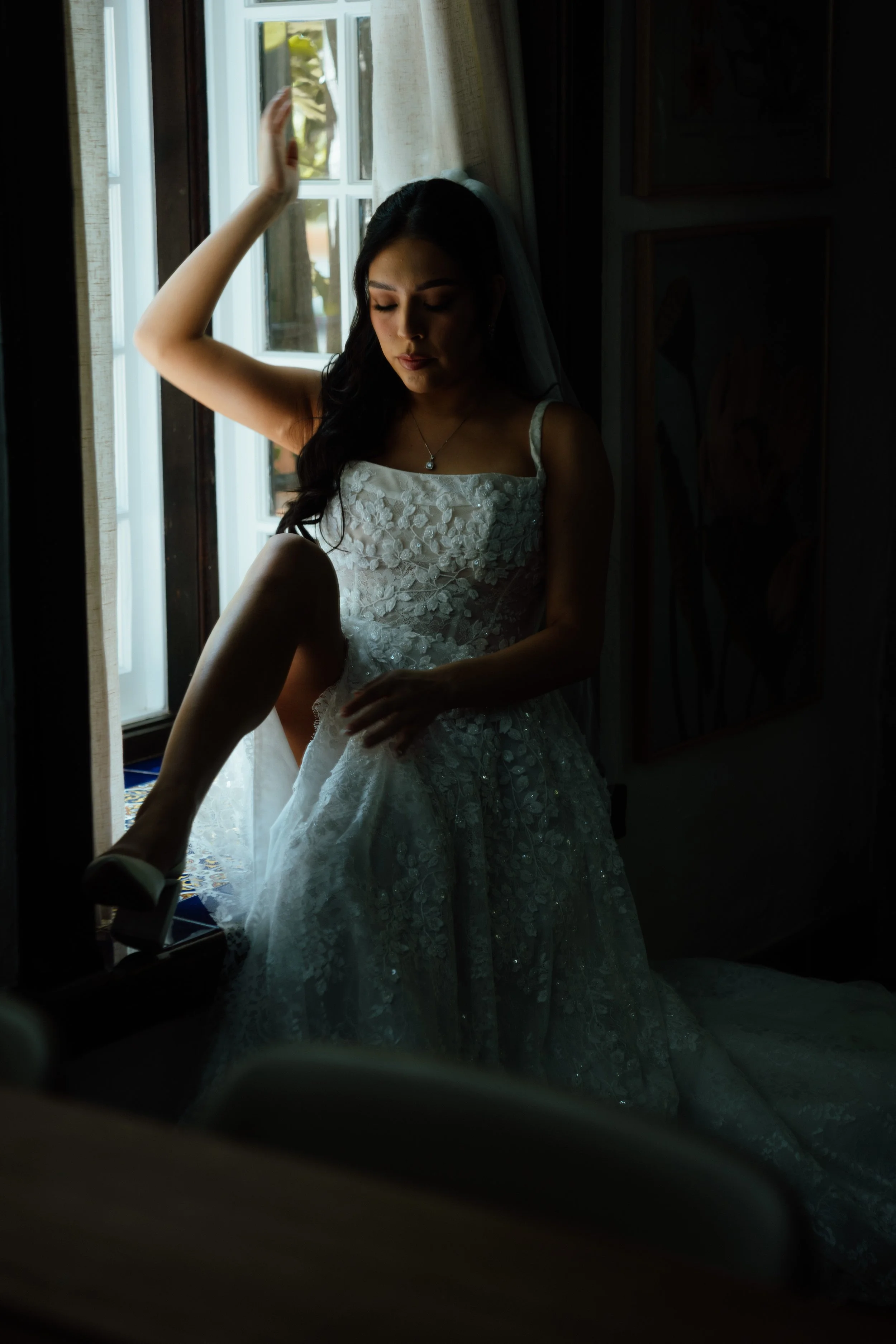 A woman in a wedding dress adjusting her shoe while sitting by a window with sunlight coming through.