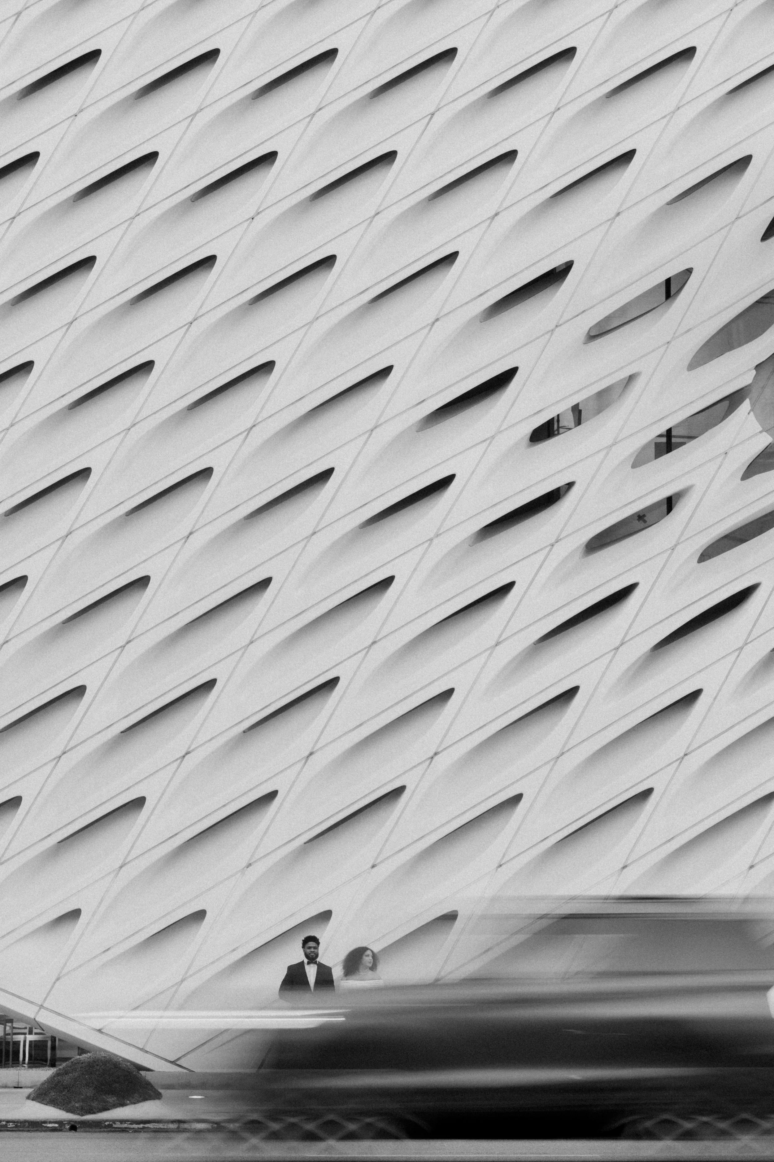 A black and white photo of an architectural structure with repeating oval-shaped openings. Two people, a man in a tuxedo and a woman in a dress, are standing inside the structure near the bottom of the image.