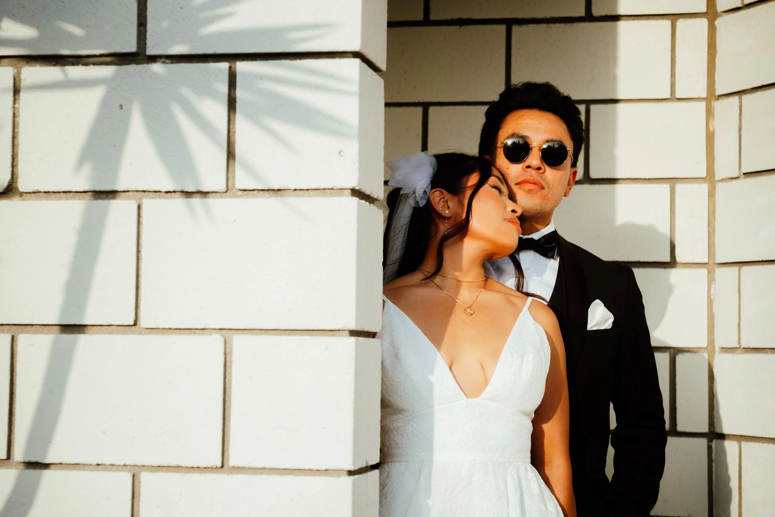 A couple dressed in wedding attire, with the woman in a white gown and the man in a black tuxedo, standing close to each other against a white brick wall. The woman is resting her head against the man's shoulder, and she is wearing a white flower hai