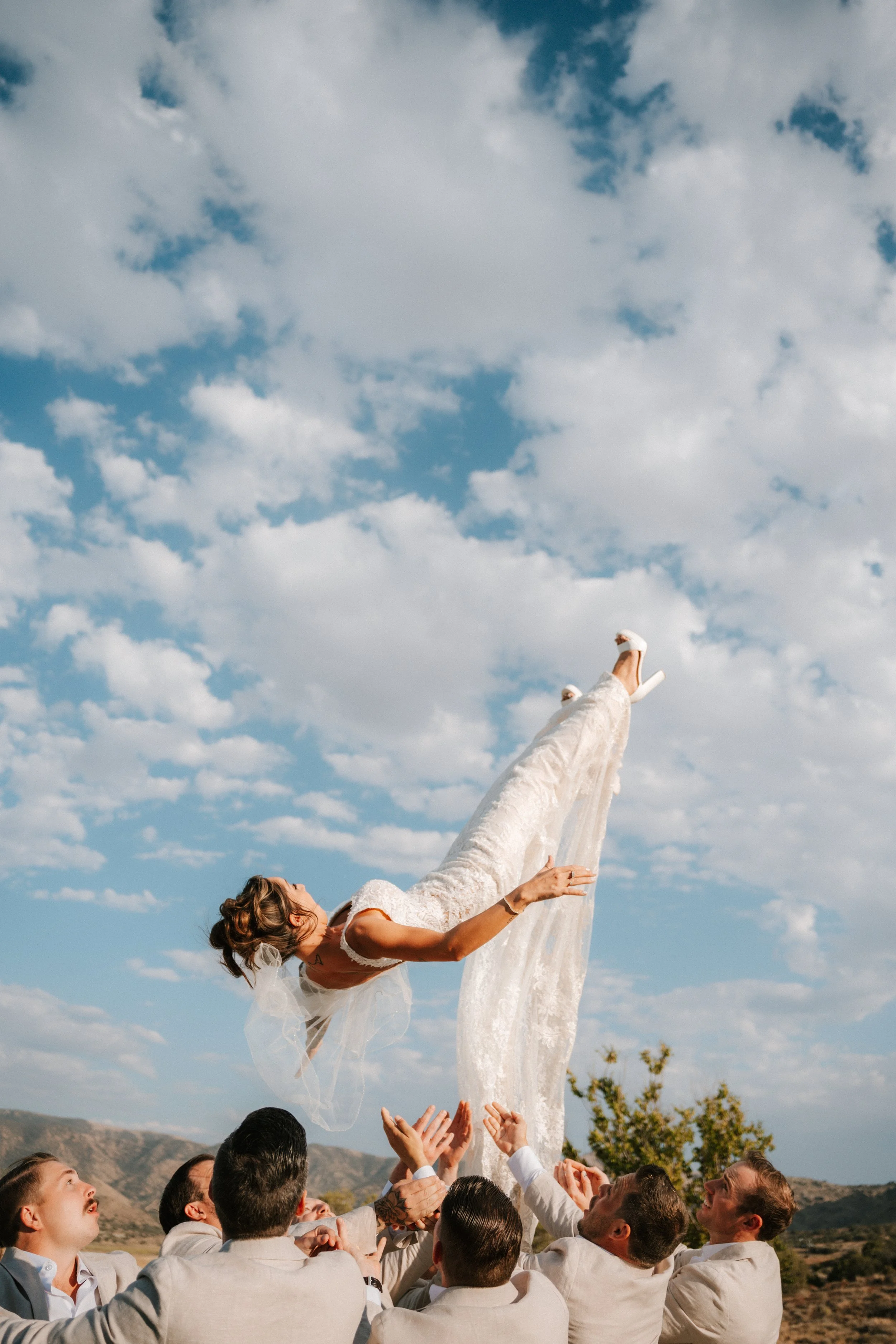A bride in a wedding dress is being lifted into the air by a group of men in suits outdoors, with mountains and trees in the background under a partly cloudy sky.