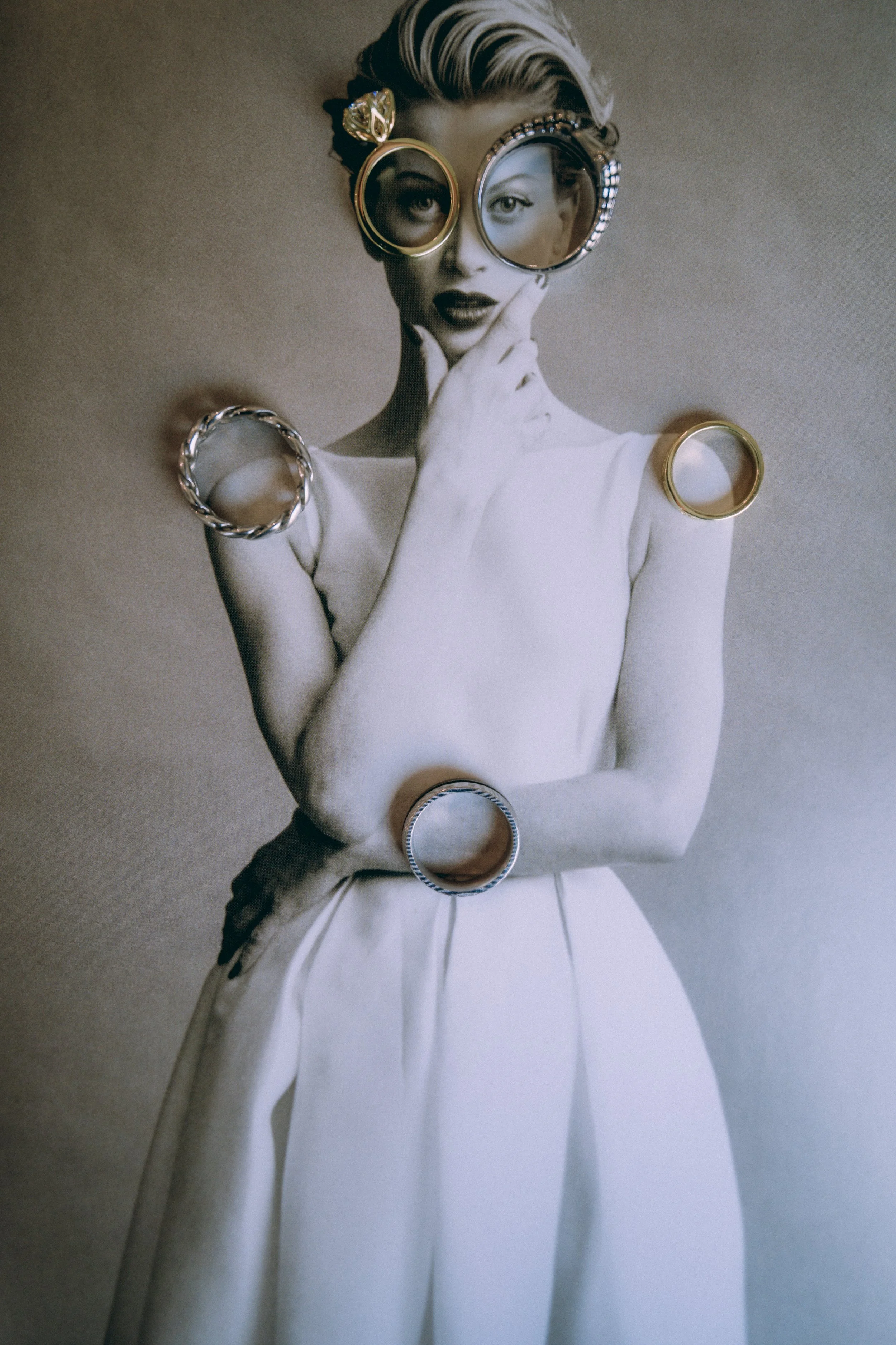A black and white photograph of a woman wearing large, round sunglasses with metallic frames. She is dressed in a white outfit, and her hair is styled with a wave. Multiple rings adorn her fingers and wrists, adding a decorative element. The woman ha
