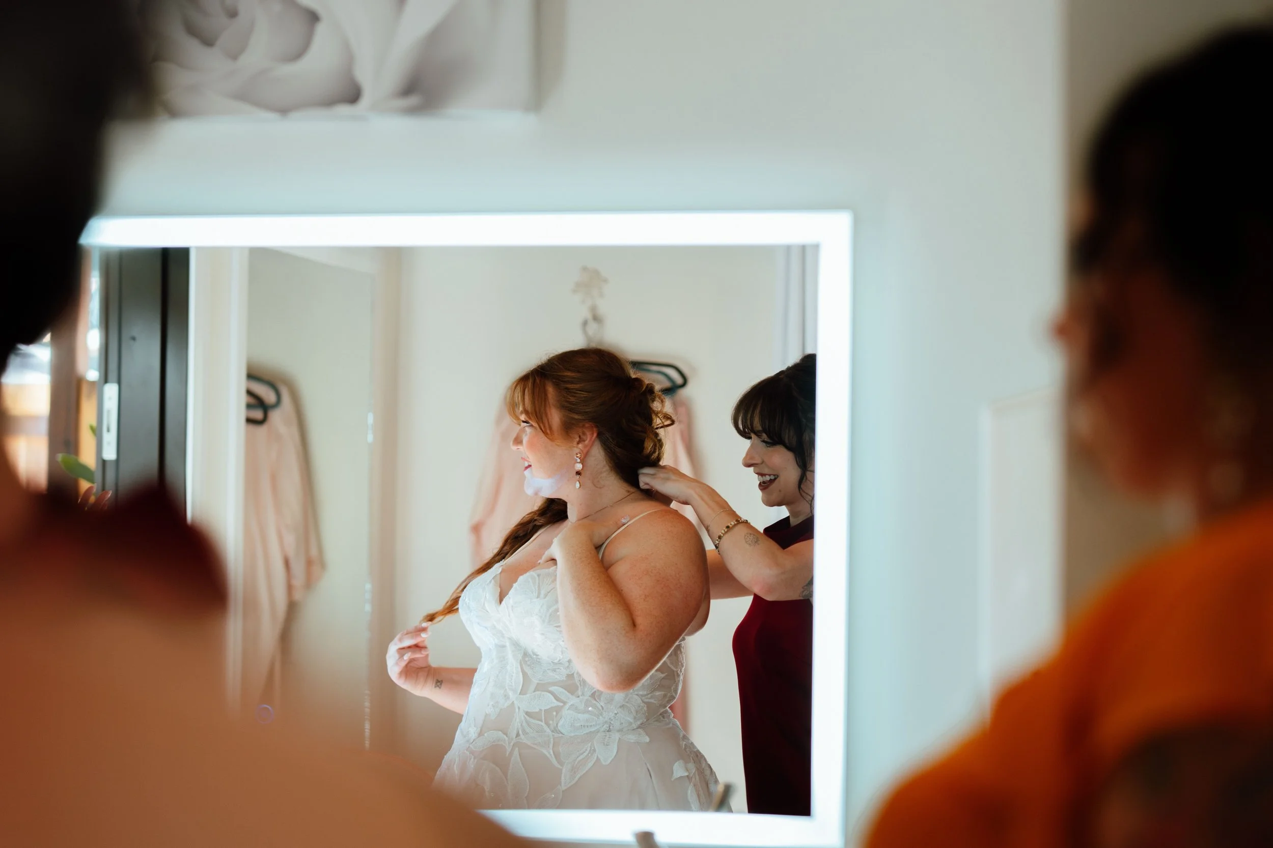 A woman in a wedding dress is being assisted by another woman in a red dress, both smiling, as they prepare in front of a mirror.