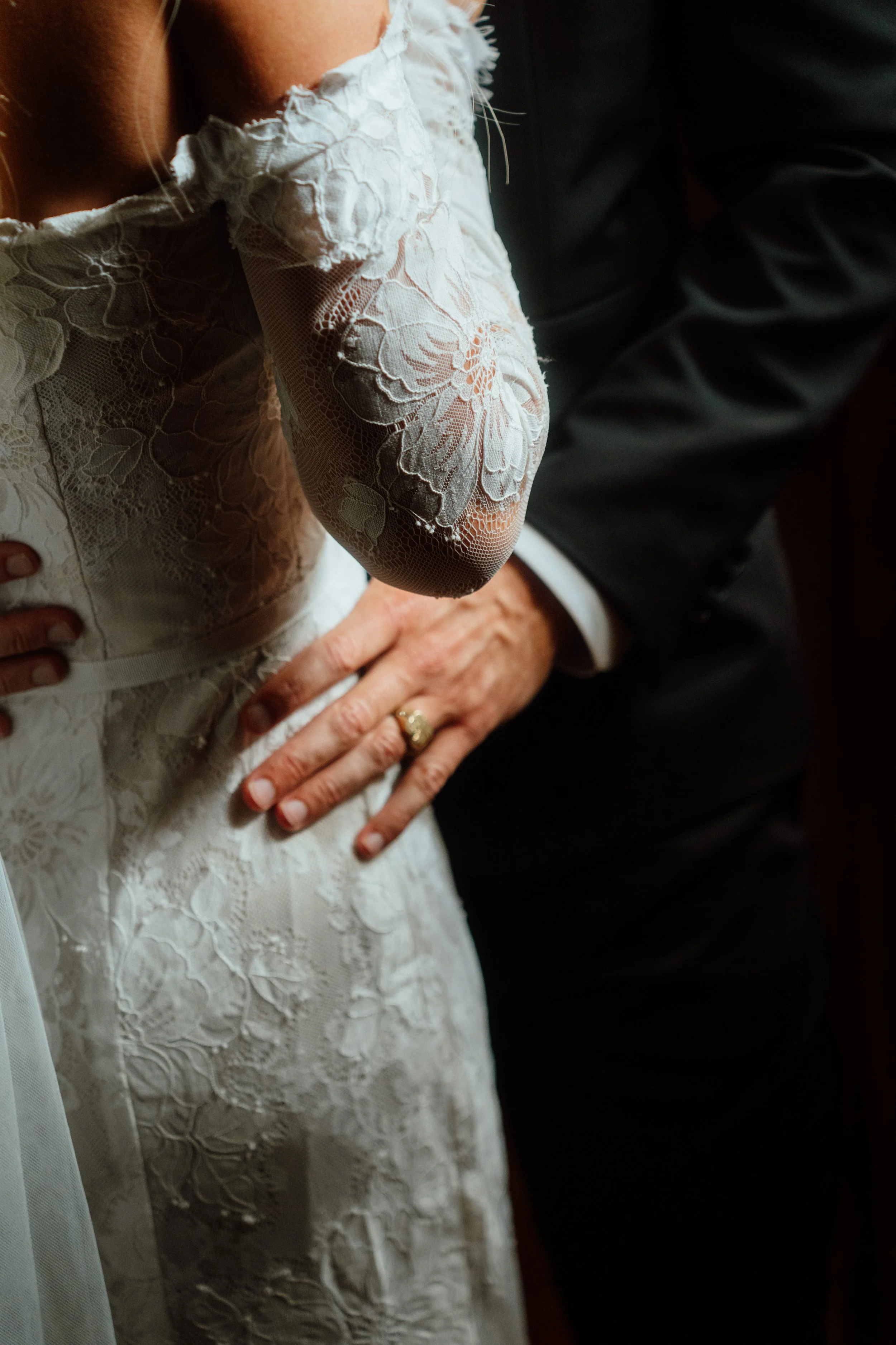 Close-up of a woman wearing a white lace dress with floral embroidery, holding her hand on her waist, and standing next to a person in a dark suit.