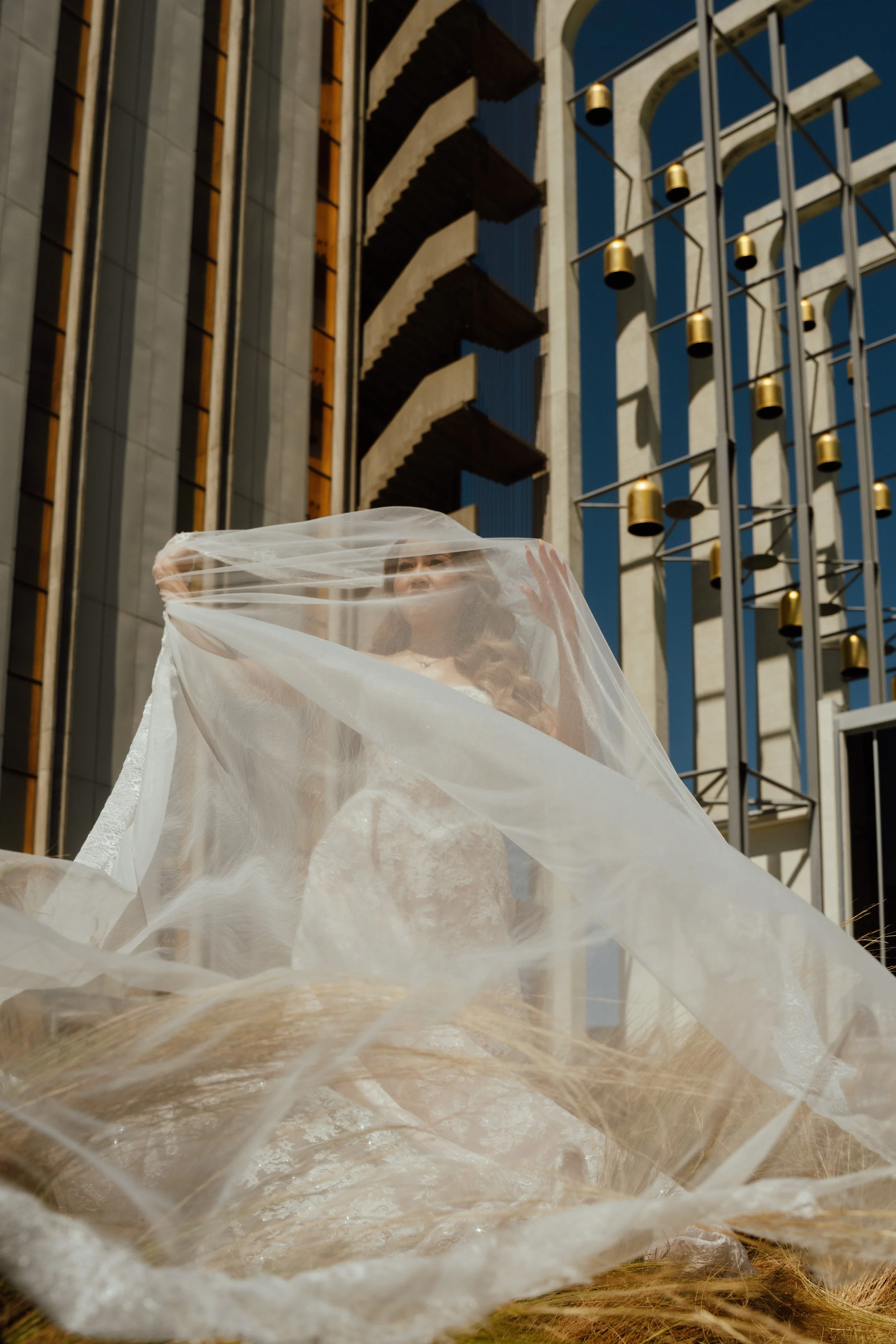 A woman in a wedding dress holding a sheer veil, standing outdoors with modern buildings and blue sky in the background.