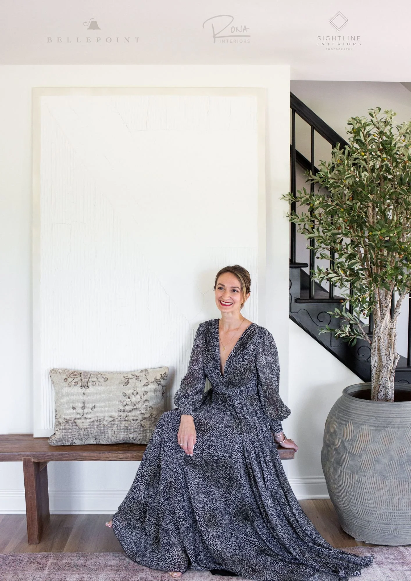 Smiling woman sitting in modern living space with large plant and decorative pillow, wearing a long patterned dress.