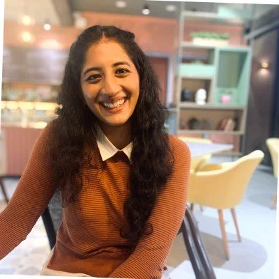 A young woman with curly dark hair is smiling and sitting at a table in a modern cafe or restaurant with pastel-colored furniture and shelves in the background.