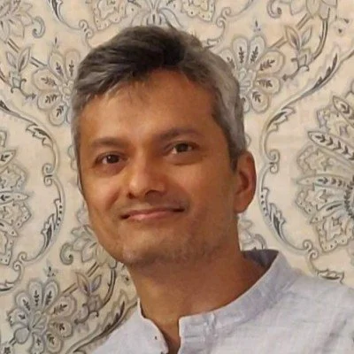 A smiling man with short gray hair, wearing a light-colored traditional shirt, standing in front of an ornate patterned background.