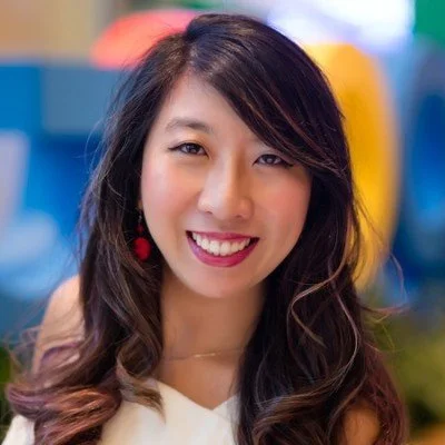 Close-up of a smiling young woman with long dark hair and earrings in front of colorful blurred background.