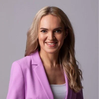 Portrait of a woman with long blonde hair wearing a pink blazer and white top, smiling against a gray background.