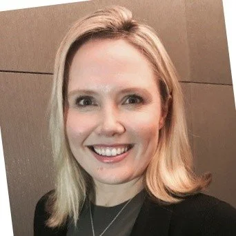 A smiling woman with blonde hair wearing a black top, standing against a wooden wall background.