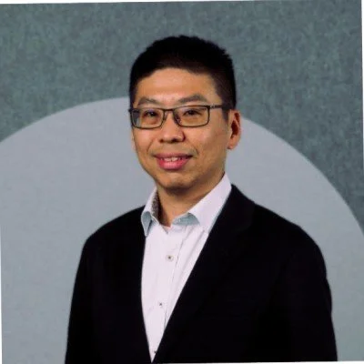 A man with black hair, glasses, and a slight smile, wearing a white shirt and a dark blazer, standing in front of a gray background with a circular design.