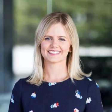 A smiling woman with blonde hair in a dark floral dress standing outdoors near a window with trees in the background.