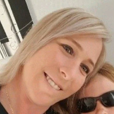 A smiling woman with blonde hair taking a selfie, with part of another person wearing sunglasses visible in the background.