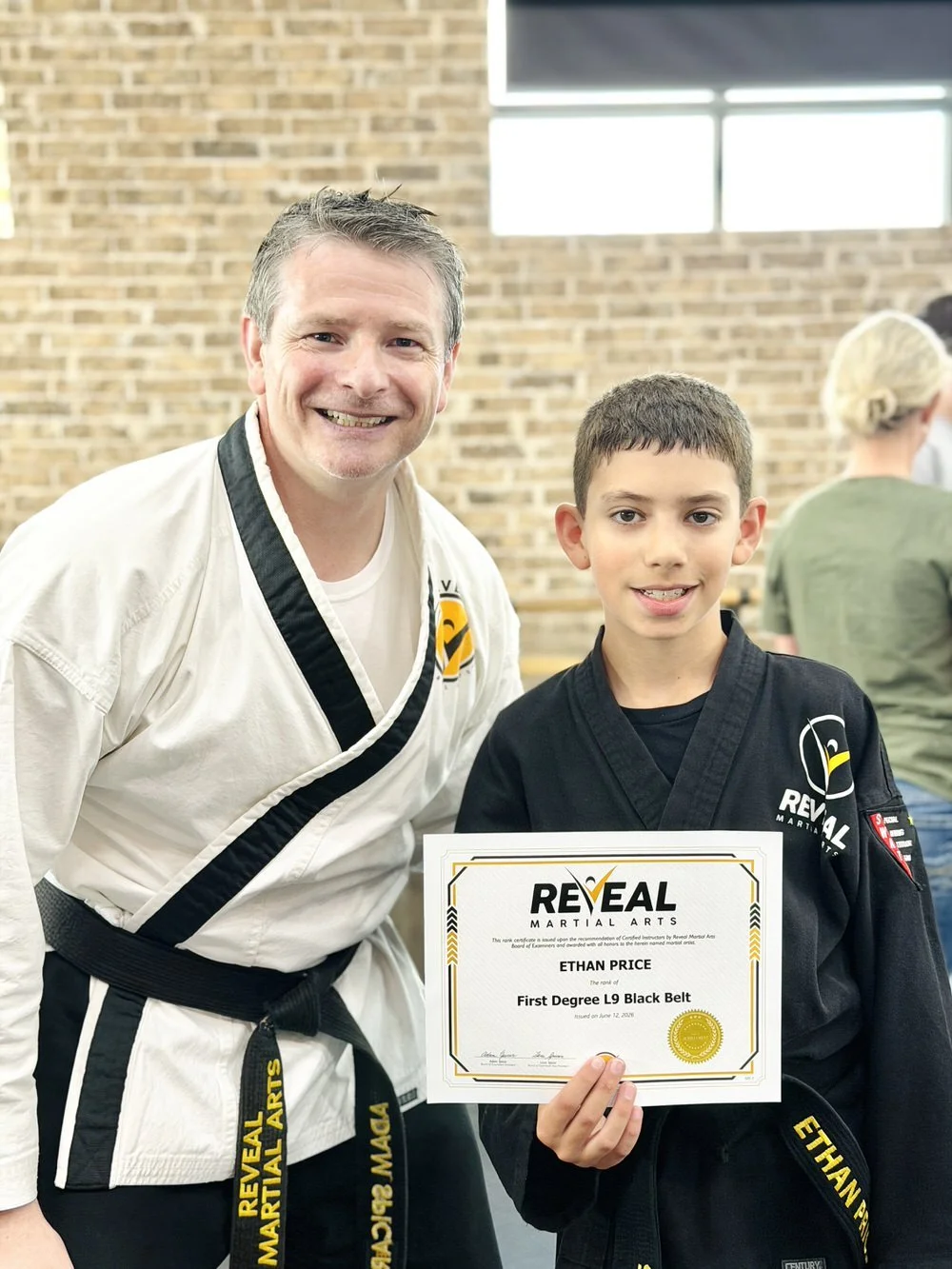 Reveal Martial Arts instructor posing with student Ethan Price holding his First Degree Black Belt certificate after promotion testing.
