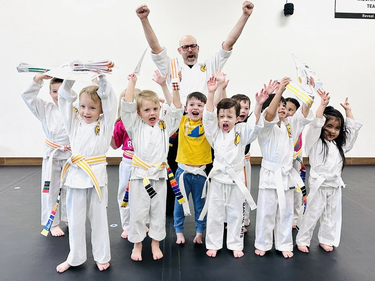 Group of kids with their Reveal Martial Arts instructor raising hand victoriously and confidently with new belts and certificates.