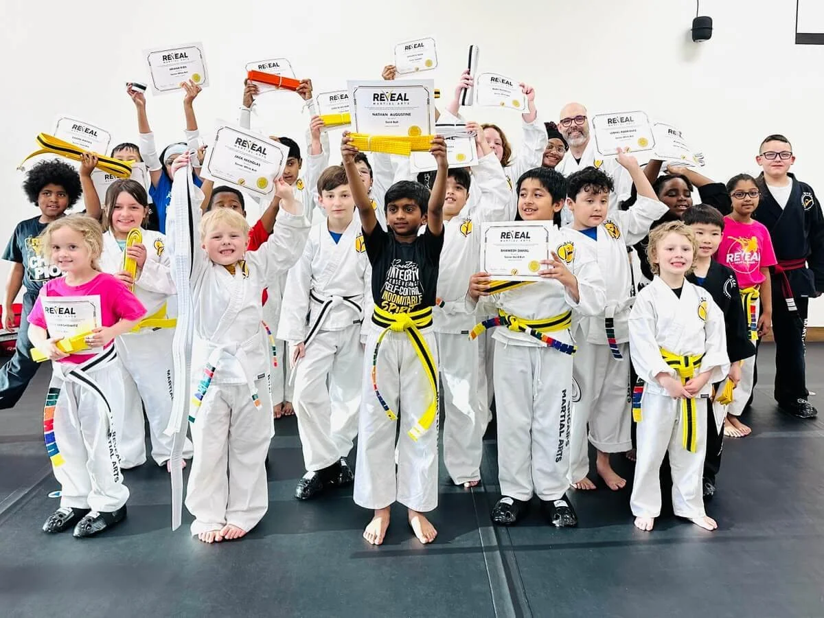 Group of children in a martial arts class victoriously holding their new belts and rank certificates high above their heads at Reveal Martial Arts.