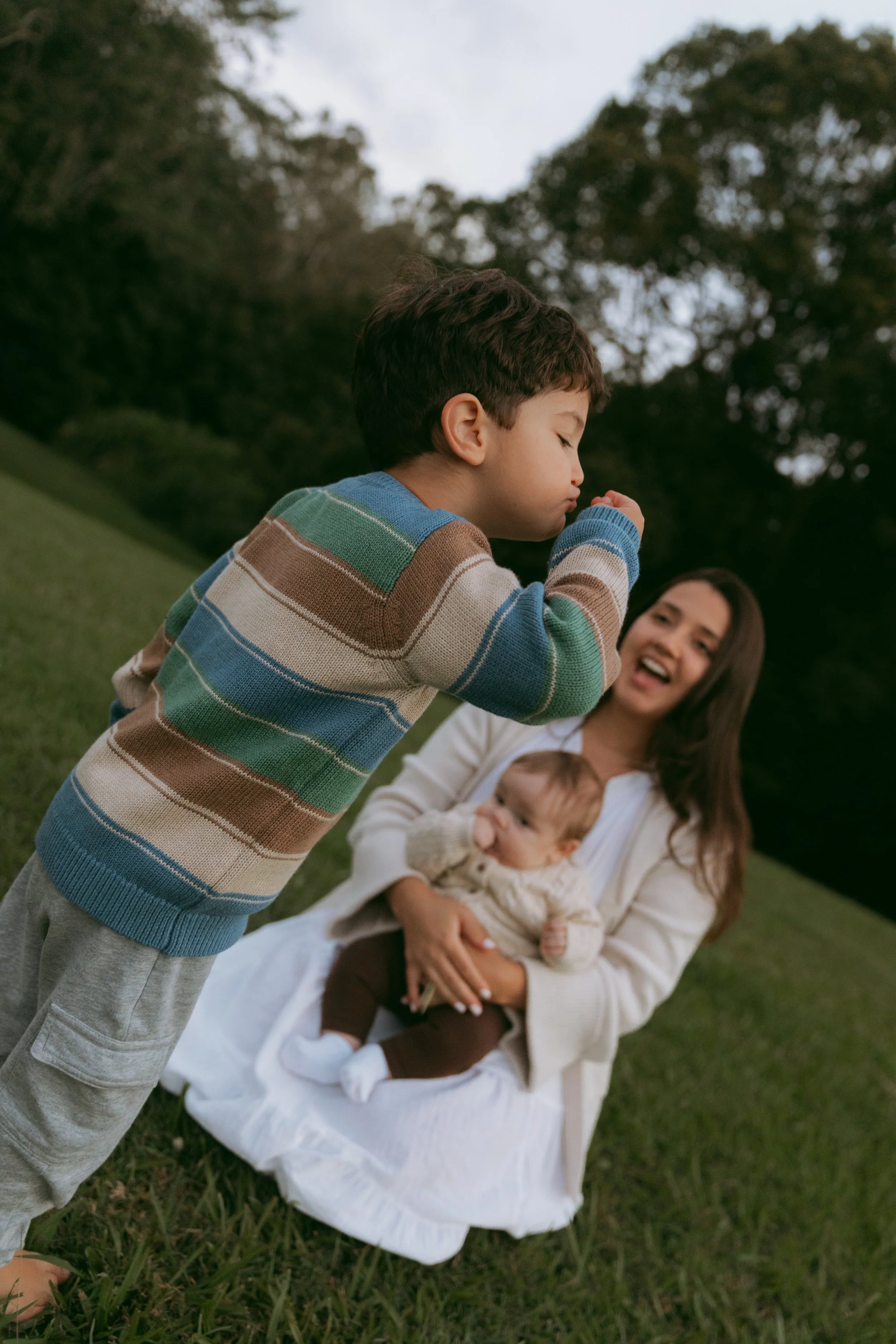 A young boy in a multicolored striped sweater stands outdoors near a woman sitting on the grass. The woman holds a baby in her lap, and the boy is blowing a bubble. There are trees and an overcast sky in the background.