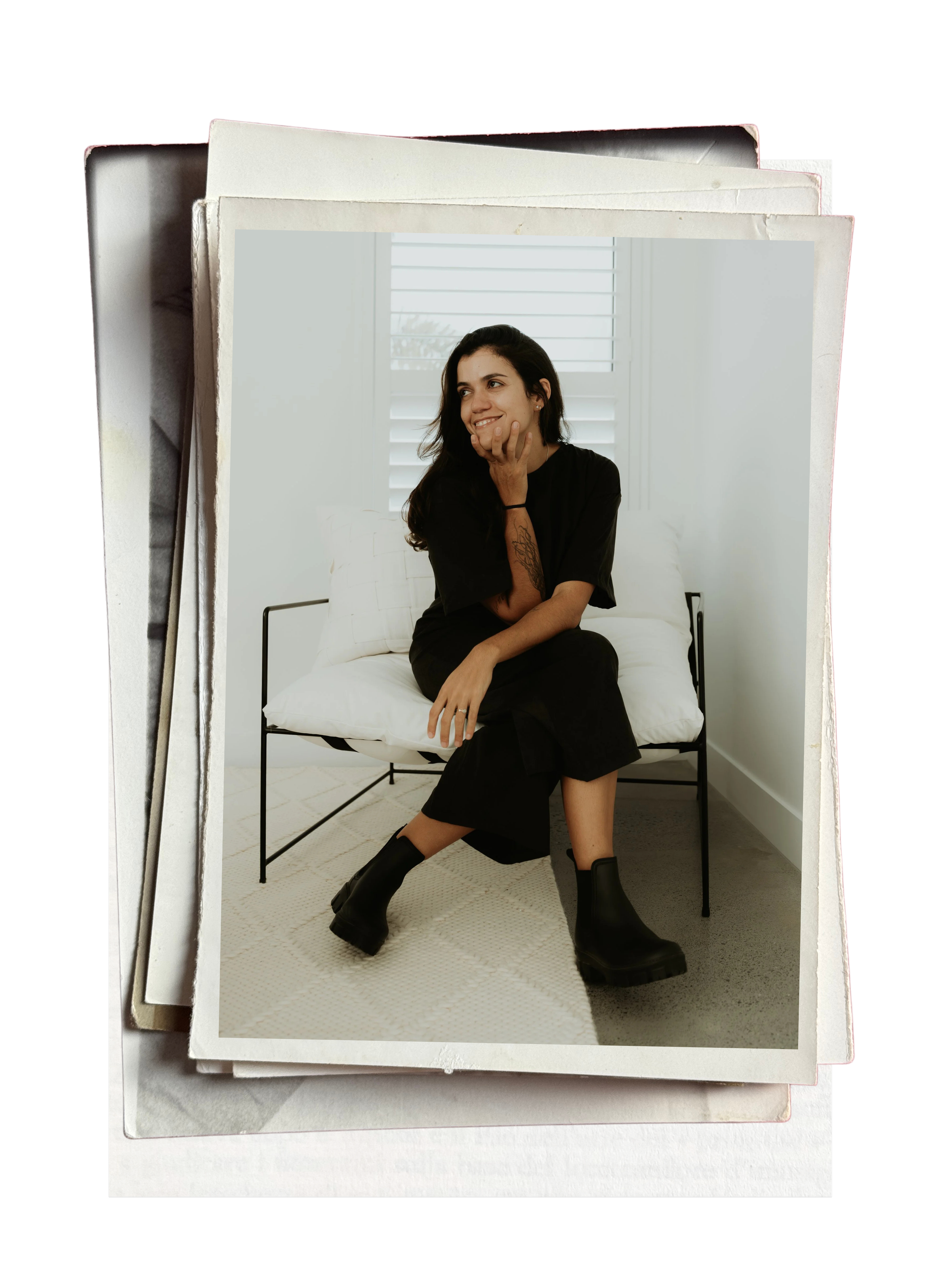 A woman with dark hair, wearing a black dress and black boots, sitting on a white bed with white pillows, smiling and resting her chin on her hand, in front of a window with white blinds.