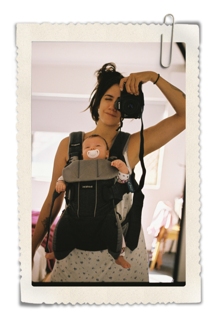 A woman taking a mirror selfie with a camera, carrying a baby in a front-facing baby carrier. The woman has dark hair, and the baby has a pacifier in their mouth.