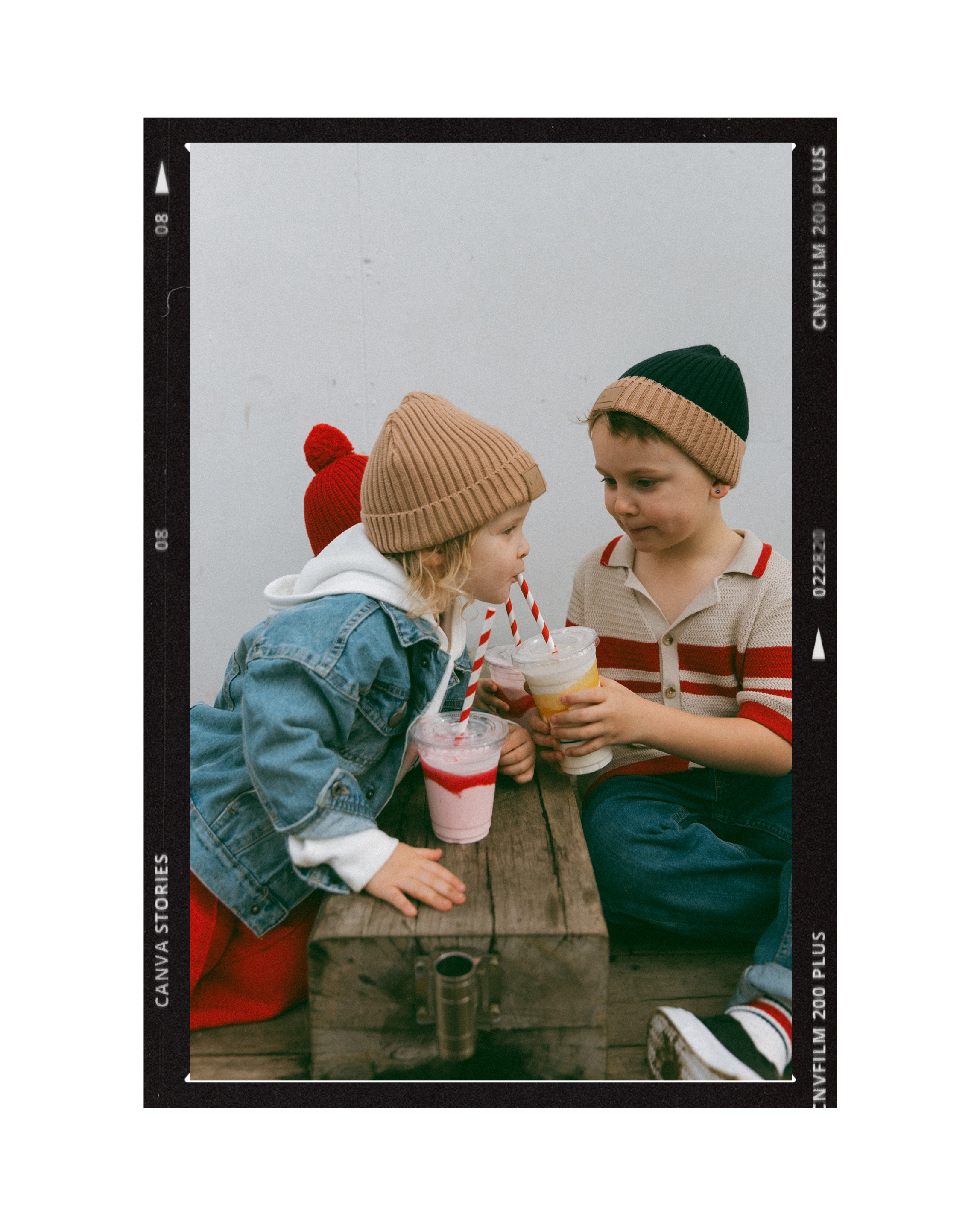 Three children sitting on a wooden picnic table, holding colorful frozen drinks with striped straws, wearing winter hats and casual clothing, outdoors against a plain background.