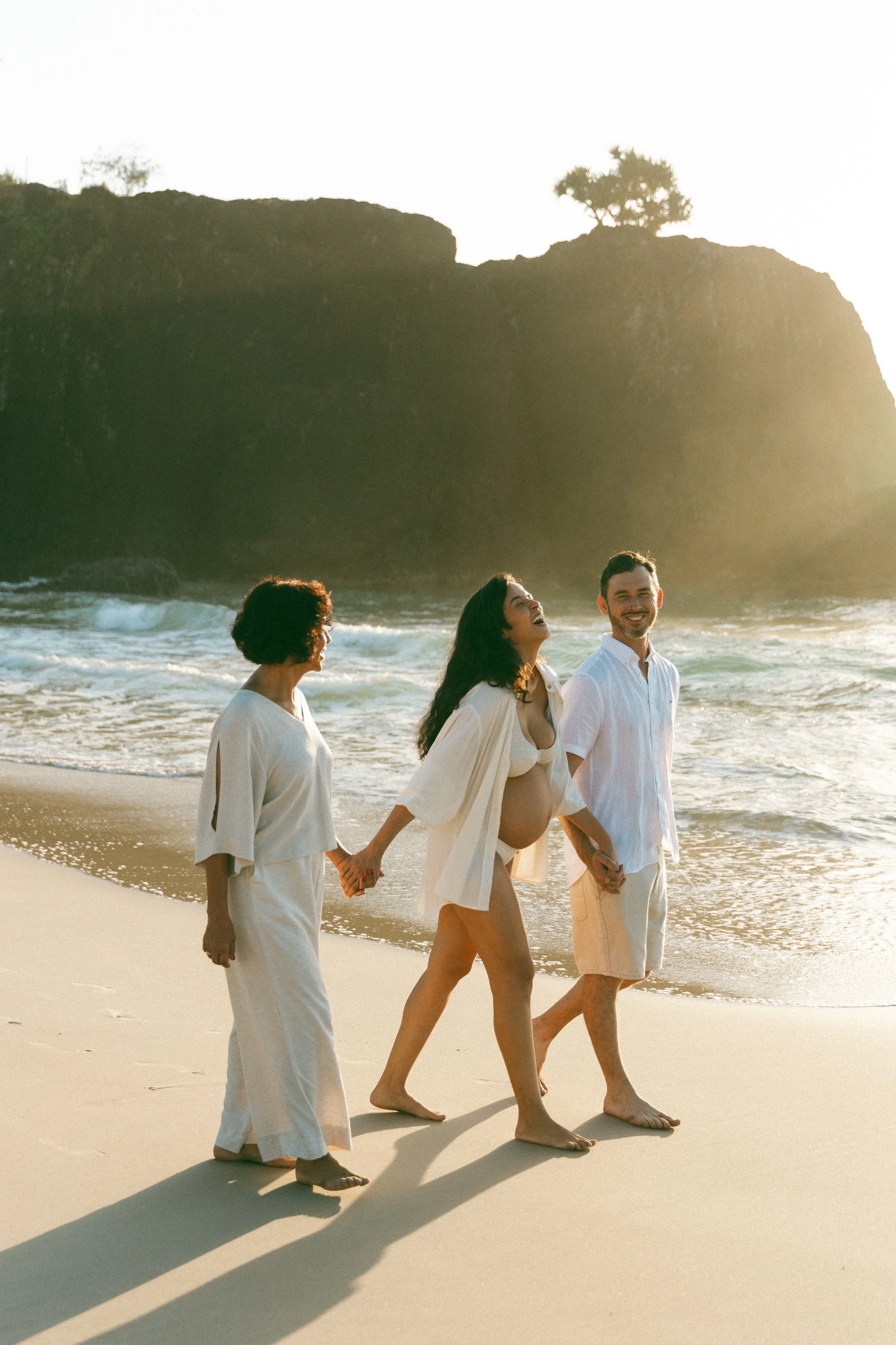 Four people walking along the beach holding hands, with a rocky cliff and a tree in the background at sunset.