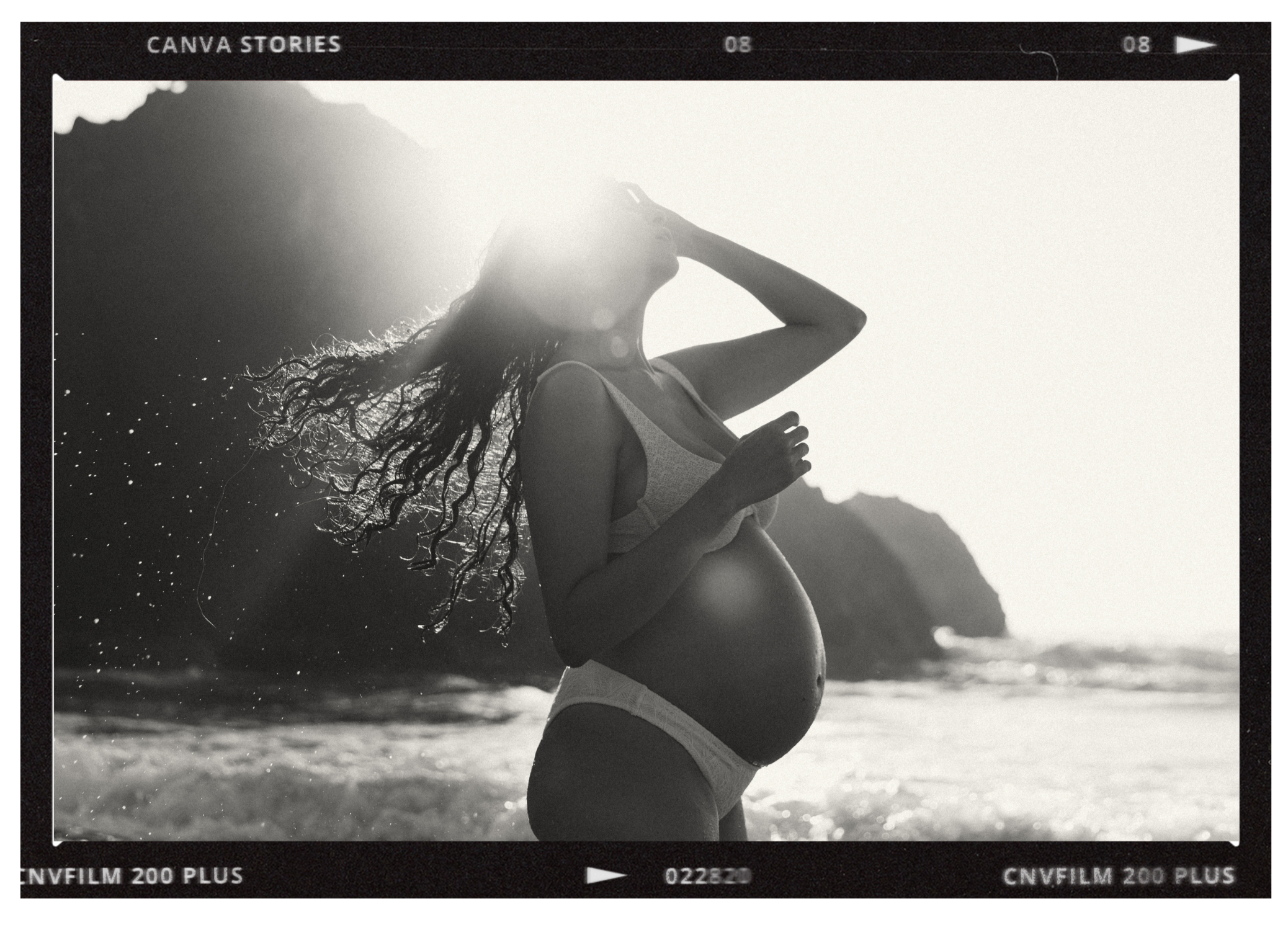 A black and white photo of a pregnant woman on the beach, standing with her hand near her face and her long curly hair blowing in the wind, with the sun shining behind her.