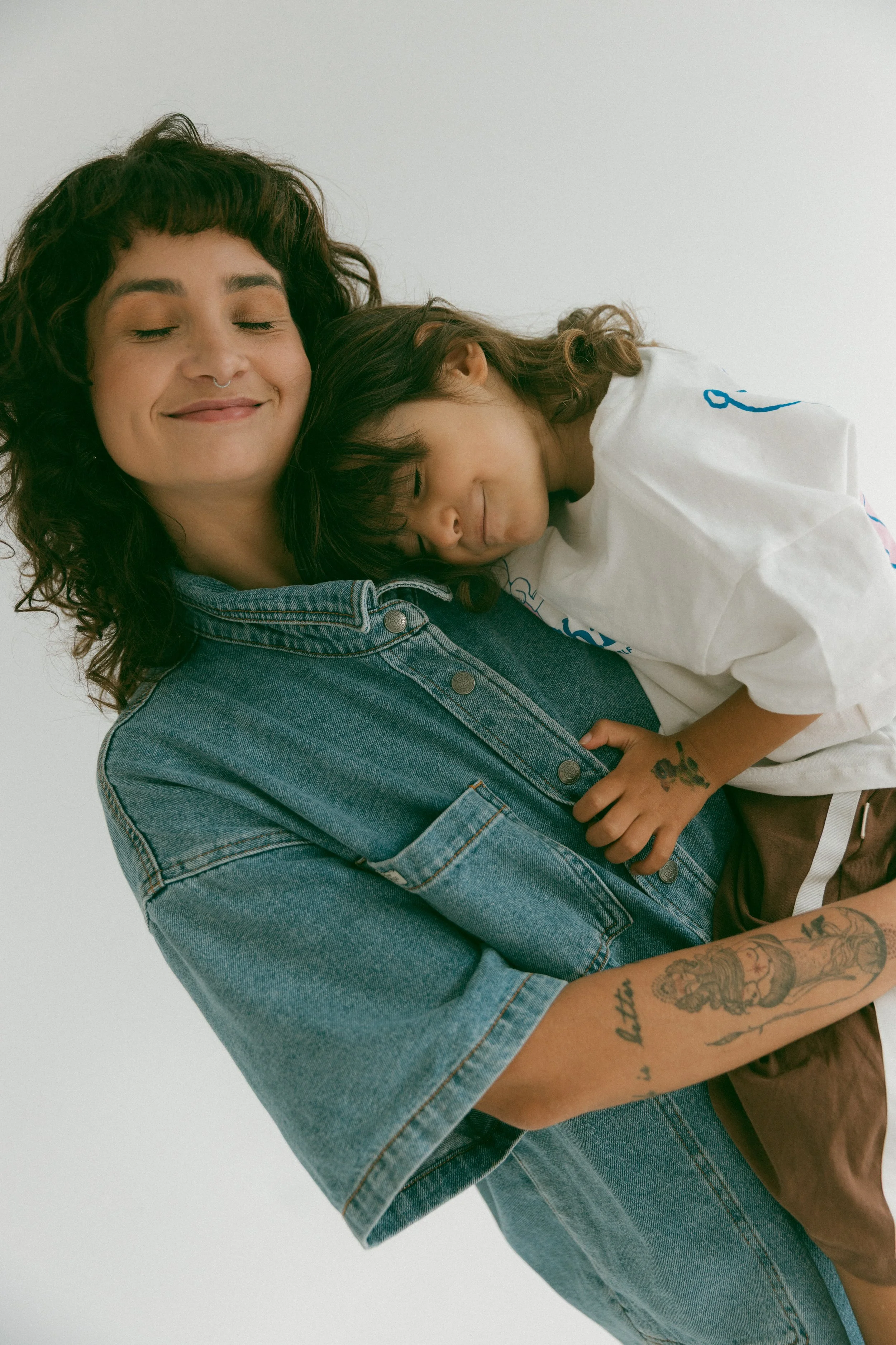 Two women, one with curly dark hair and tattoos, and one with straight brown hair and a nose ring, are hugging and smiling with their eyes closed against a plain white background.