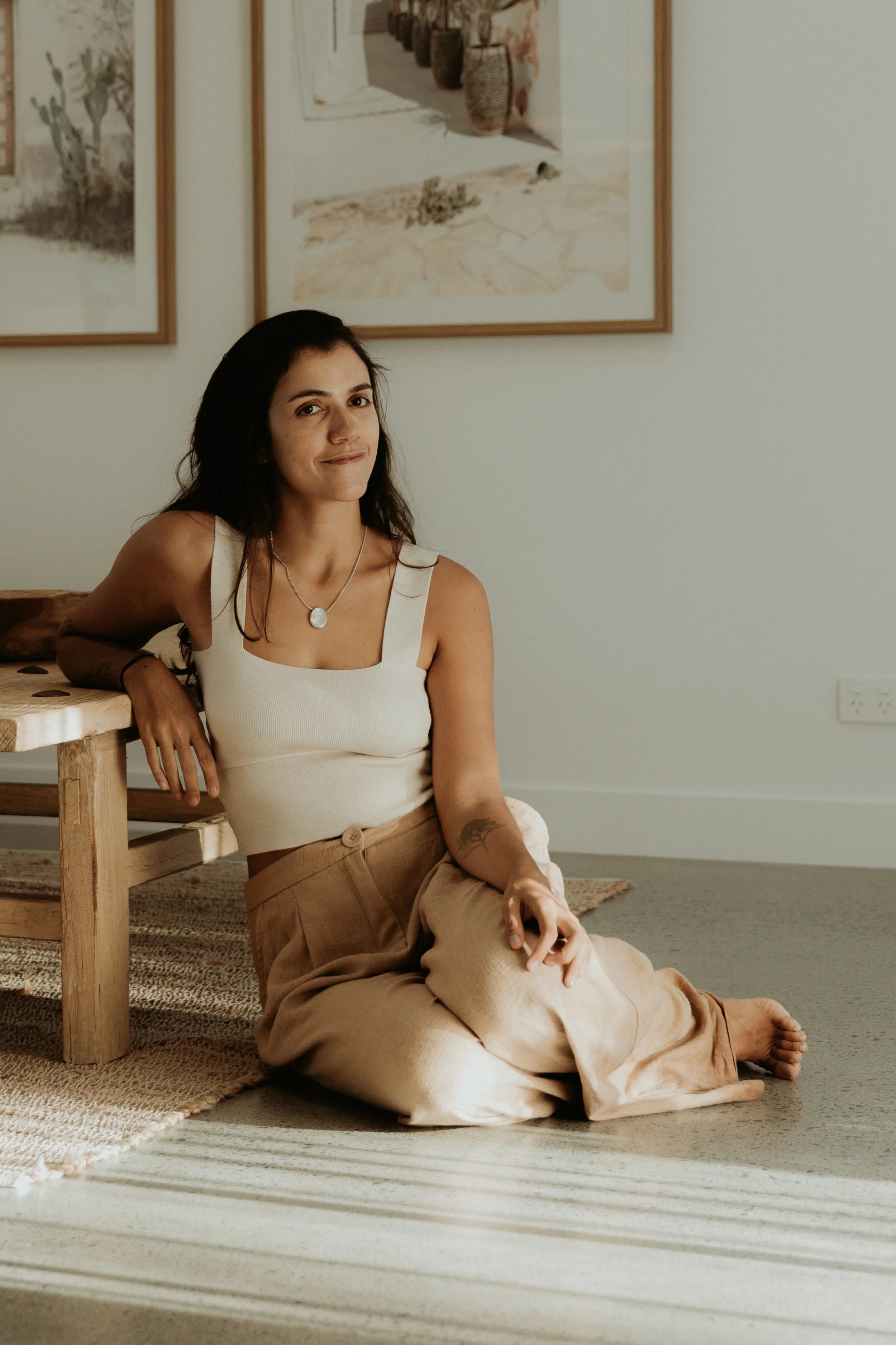A young woman with dark hair and light skin is sitting on the floor beside a wooden table, wearing a white tank top and beige pants. She is seated with one knee up, resting her arm on the table, and looking at the camera with a slight smile. The room has a white wall decorated with framed artwork, a woven rug on the floor, and sunlight creating striped shadows across the scene.