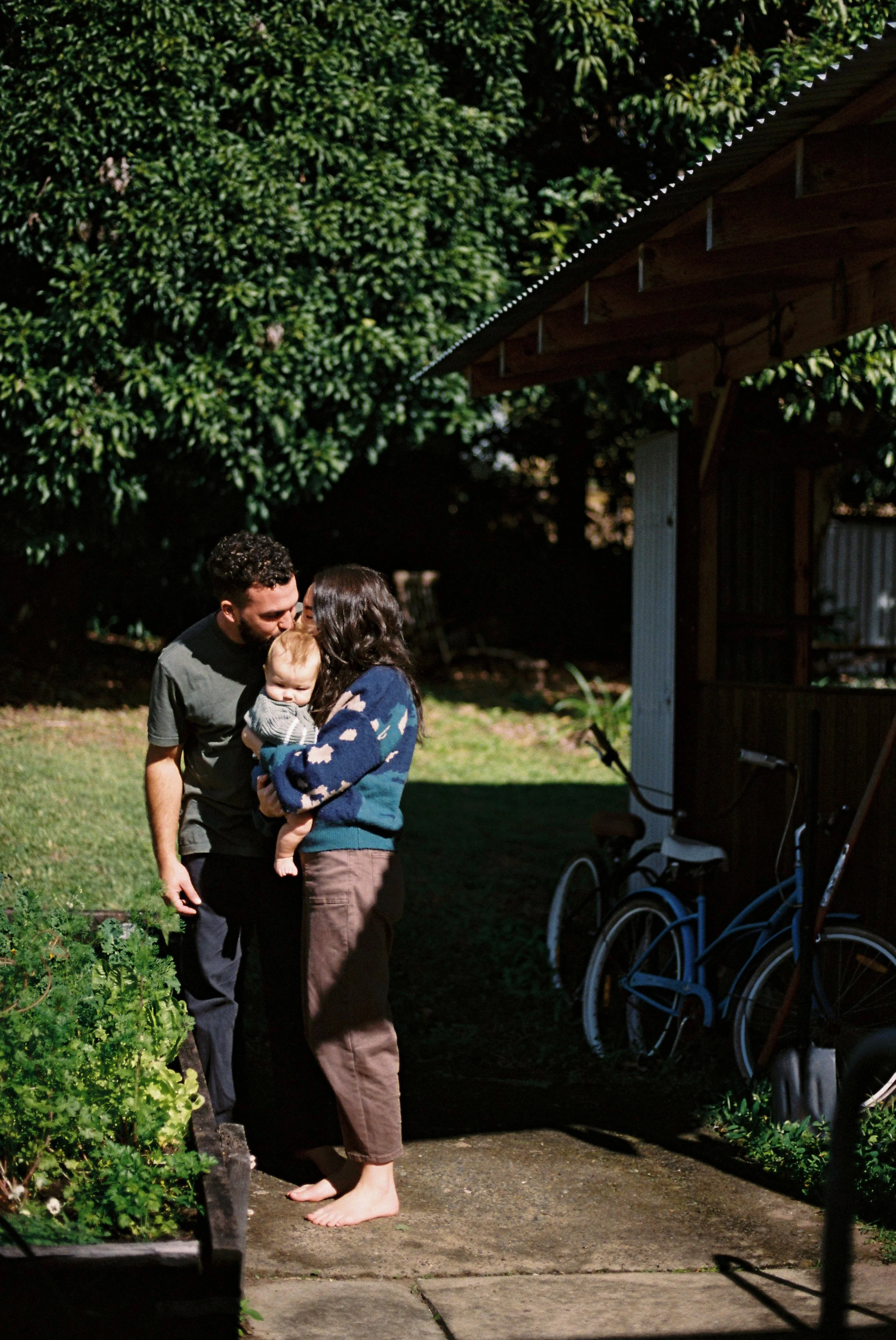 A family of three, including a man, a woman, and a young child, standing barefoot on a garden path with lush green bushes and a wooden shed in the background, during daylight hours.