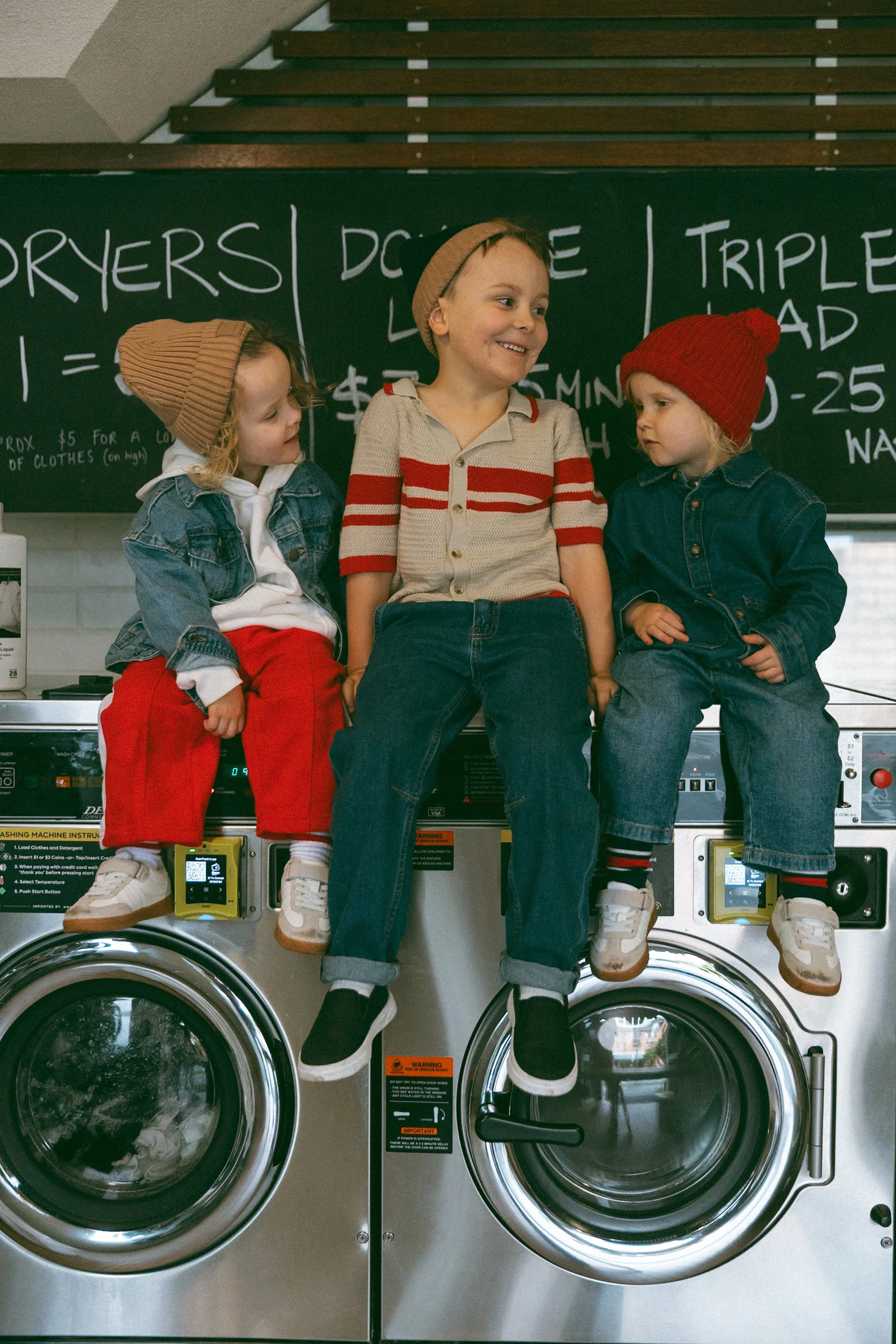 Three children sitting on top of washing machines in a laundromat. They are all smiling and wearing colorful knit hats and casual clothes.