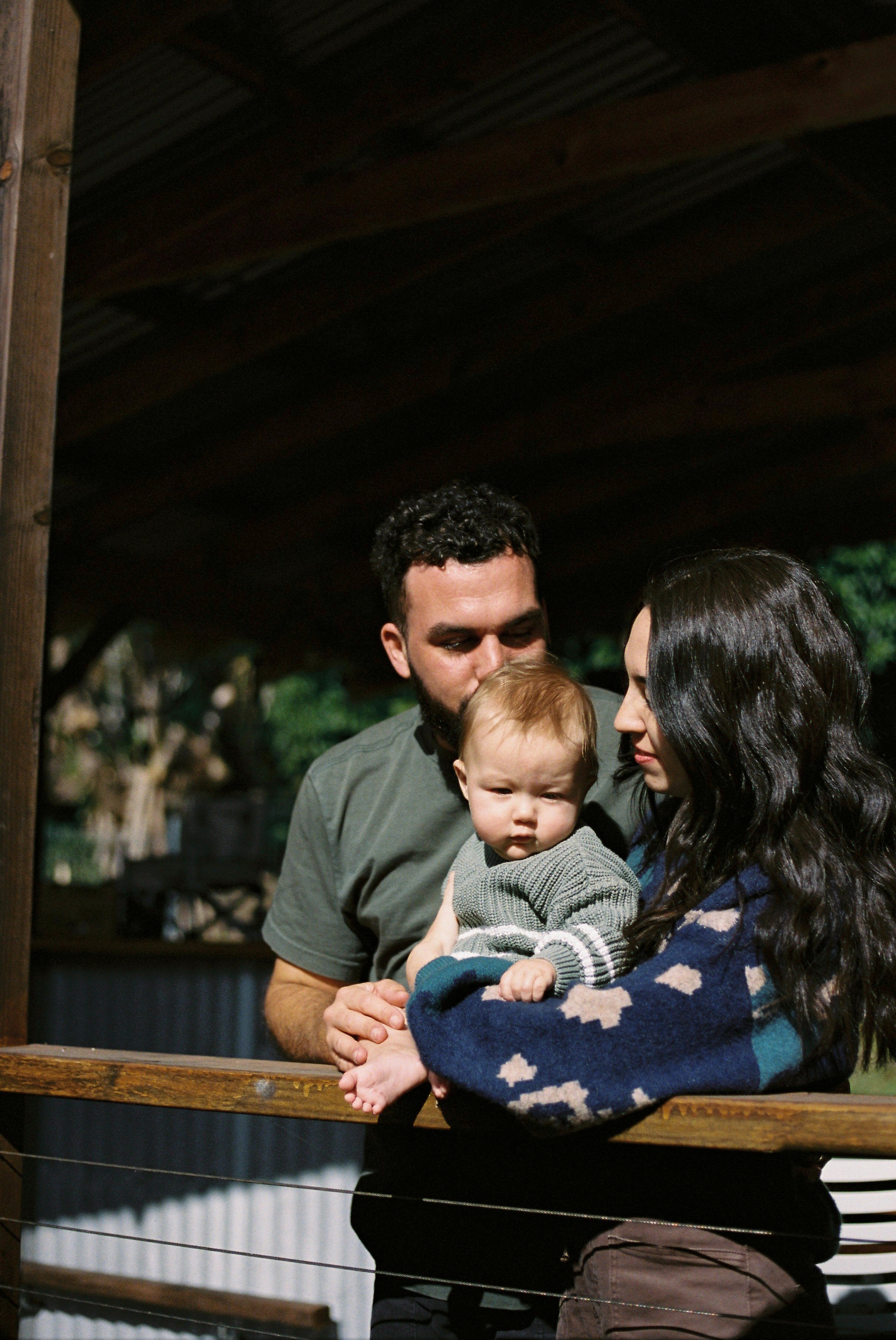 A family of three, a man, woman, and a young child, are standing close together outdoors under a wooden roof. The man is holding the child, who is looking directly at the camera, while the woman looks at the child. The setting appears to be a farm or rural area with trees in the background.
