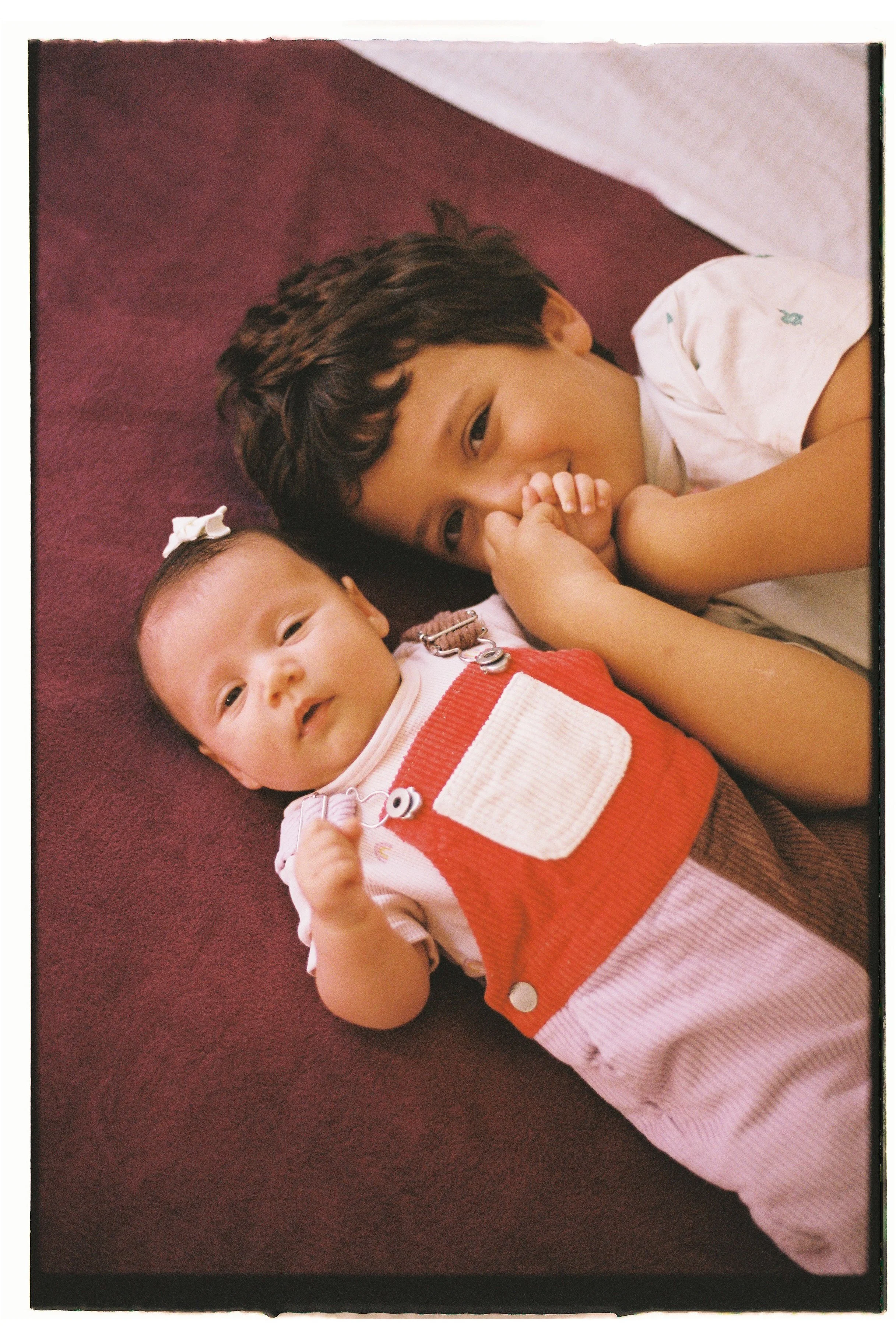 A young boy and a baby lying on a maroon surface. The boy is resting his head on the surface and looking at the camera, while the baby is lying down and looking with interest. The boy is wearing a white shirt, and the baby is dressed in a red and white outfit with buttons.