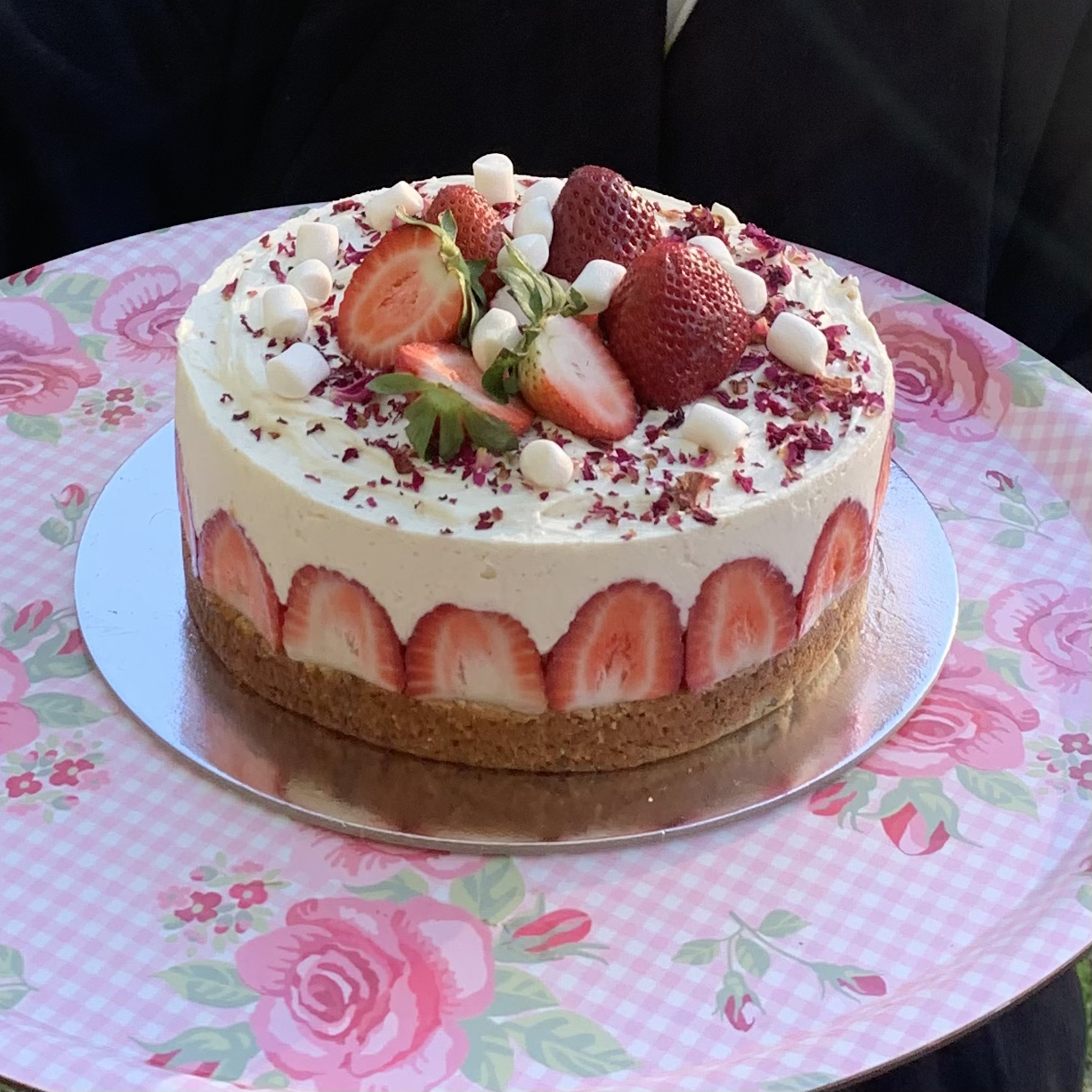 A round strawberry cake with a layer of whole strawberries around the sides, topped with sliced strawberries, white candies, and pink flower petals on a floral pink tablecloth.