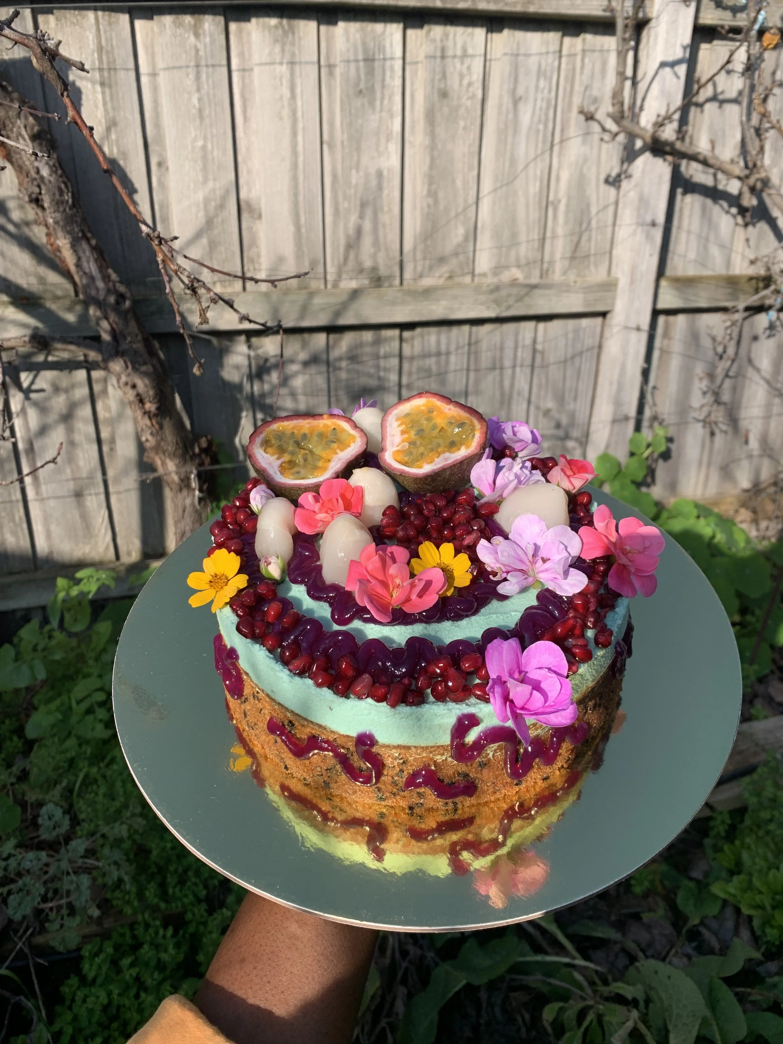 Colorful layered cake decorated with passion fruit halves, pink and purple flowers, pomegranate seeds, and white candies on top, held outdoors against a wooden fence background.