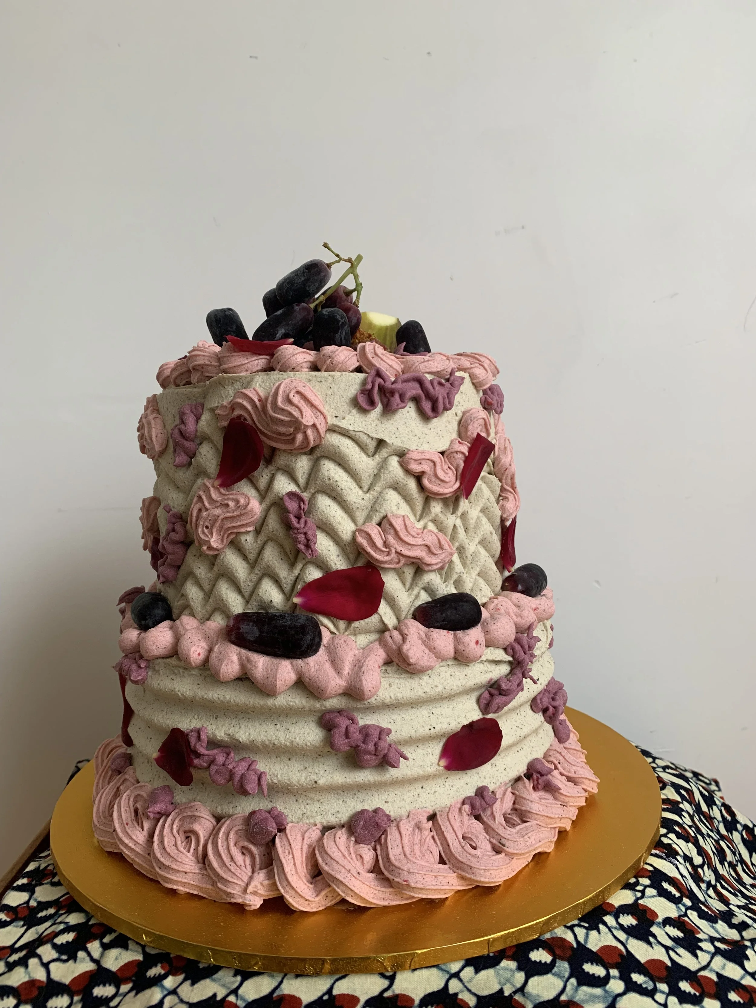 Two-tiered cake decorated with pink and purple frosting, grape cluster, and red flower petals on top, sitting on a gold cake board against a plain white background.