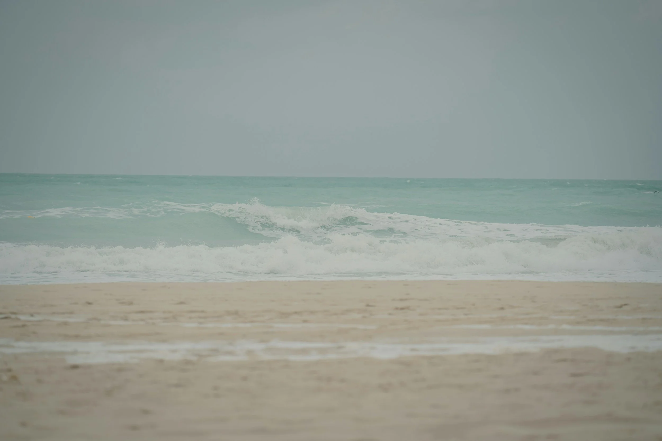 Pale turquoise sea with a wave rolling into sandy shore in muted colours
