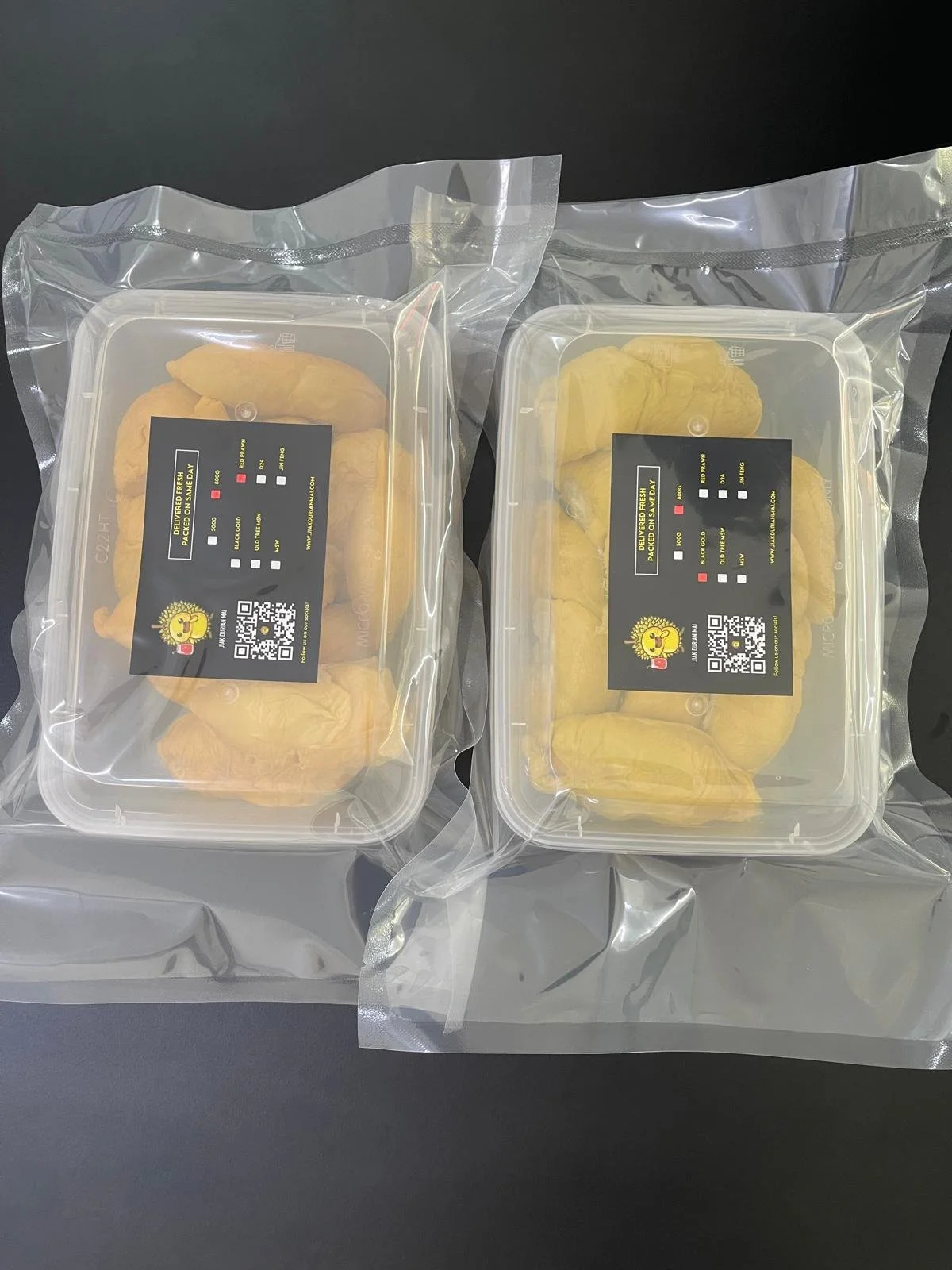 Two plastic containers of yellow durian fruit, sealed with clear plastic wrap, placed on a black surface.