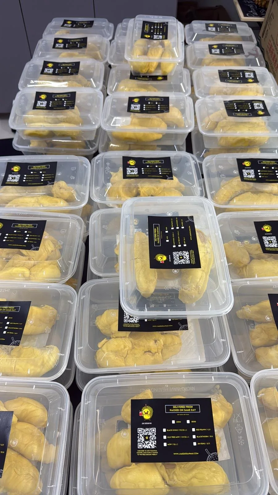 Multiple transparent plastic containers filled with durian fruit, each labeled with a black sticker that includes a QR code, brand logo, and product information.