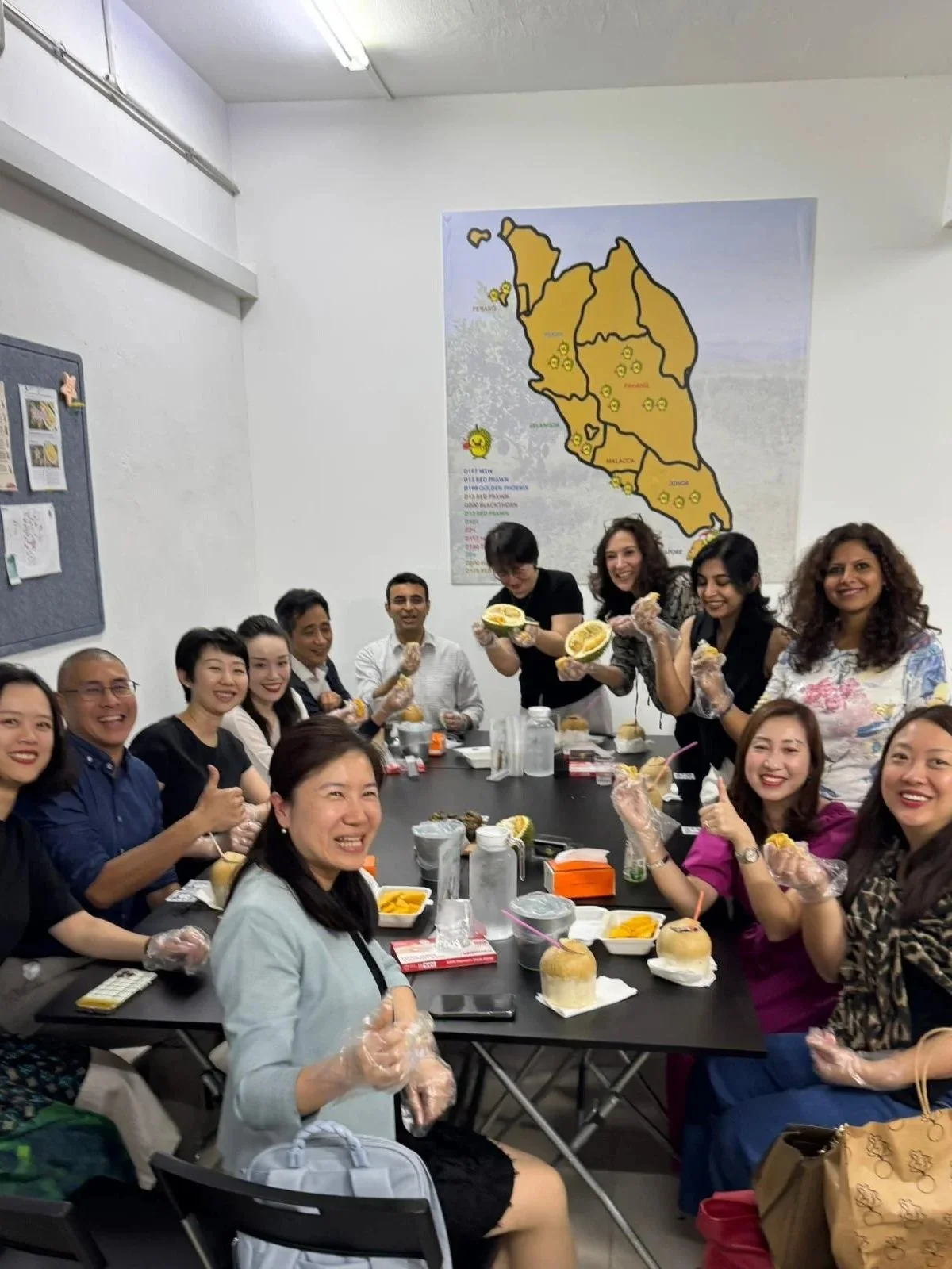 A group of diverse people sitting around a table, smiling and holding traditional Thai desserts, with a map of Thailand on the wall behind them.